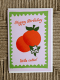 A square greeting card with a white background and a green scalloped border, featuring two orange fruits with green leaves and the text 'Happy Birthday little cutie!' in orange. The card is photographed on a textured brown woven surface.