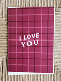 A square greeting card with a dark red and pink plaid pattern and the words 'I LOVE YOU' in white, placed on a woven straw surface.