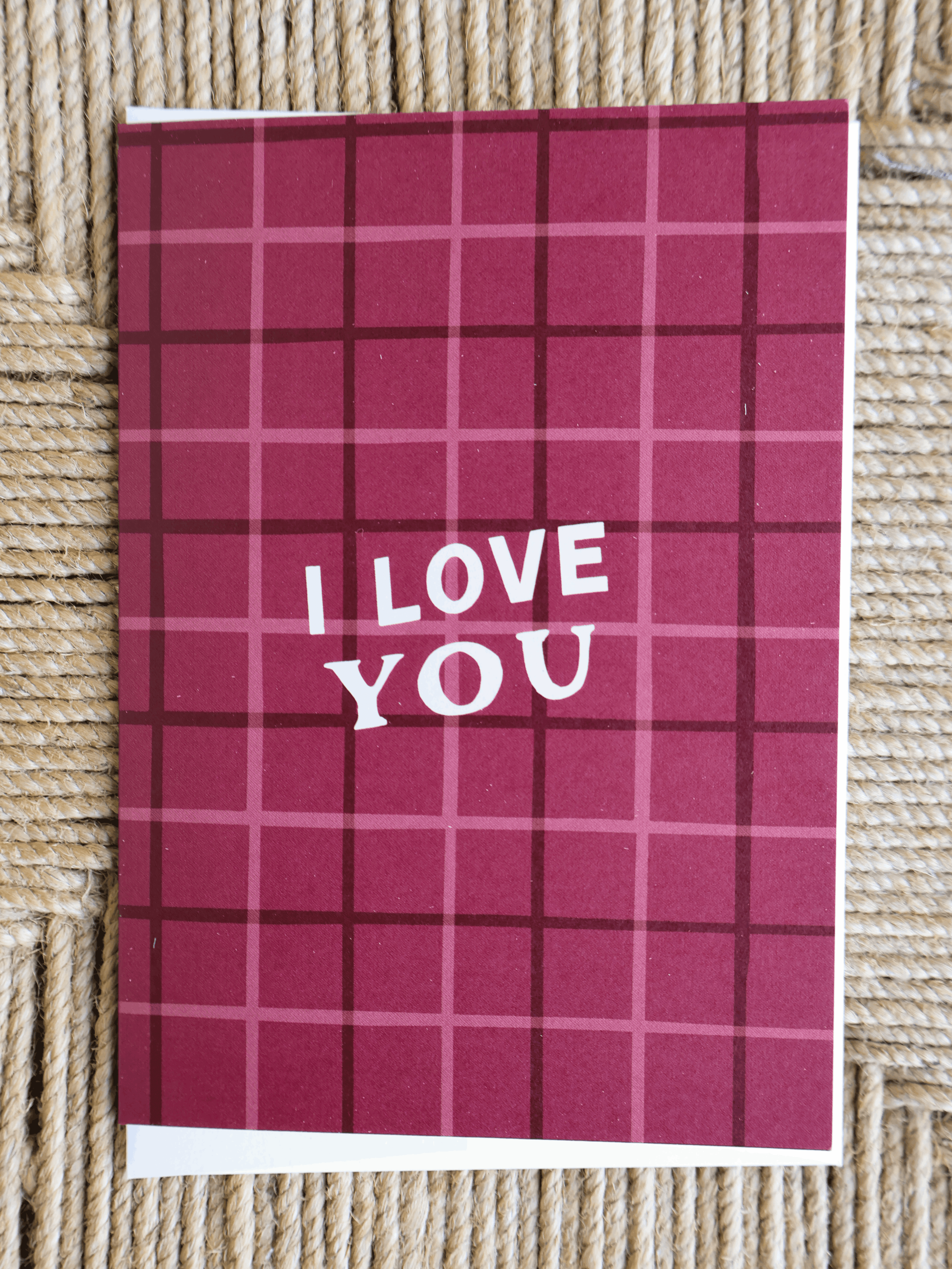 A square greeting card with a dark red and pink plaid pattern and the words 'I LOVE YOU' in white, placed on a woven straw surface.