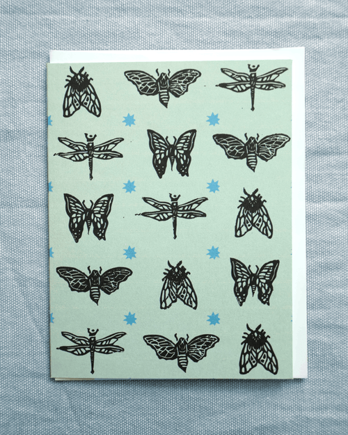 Card with black insect illustrations on a light green background with blue stars, on a textured gray surface. based on original linocut designs with envelope included