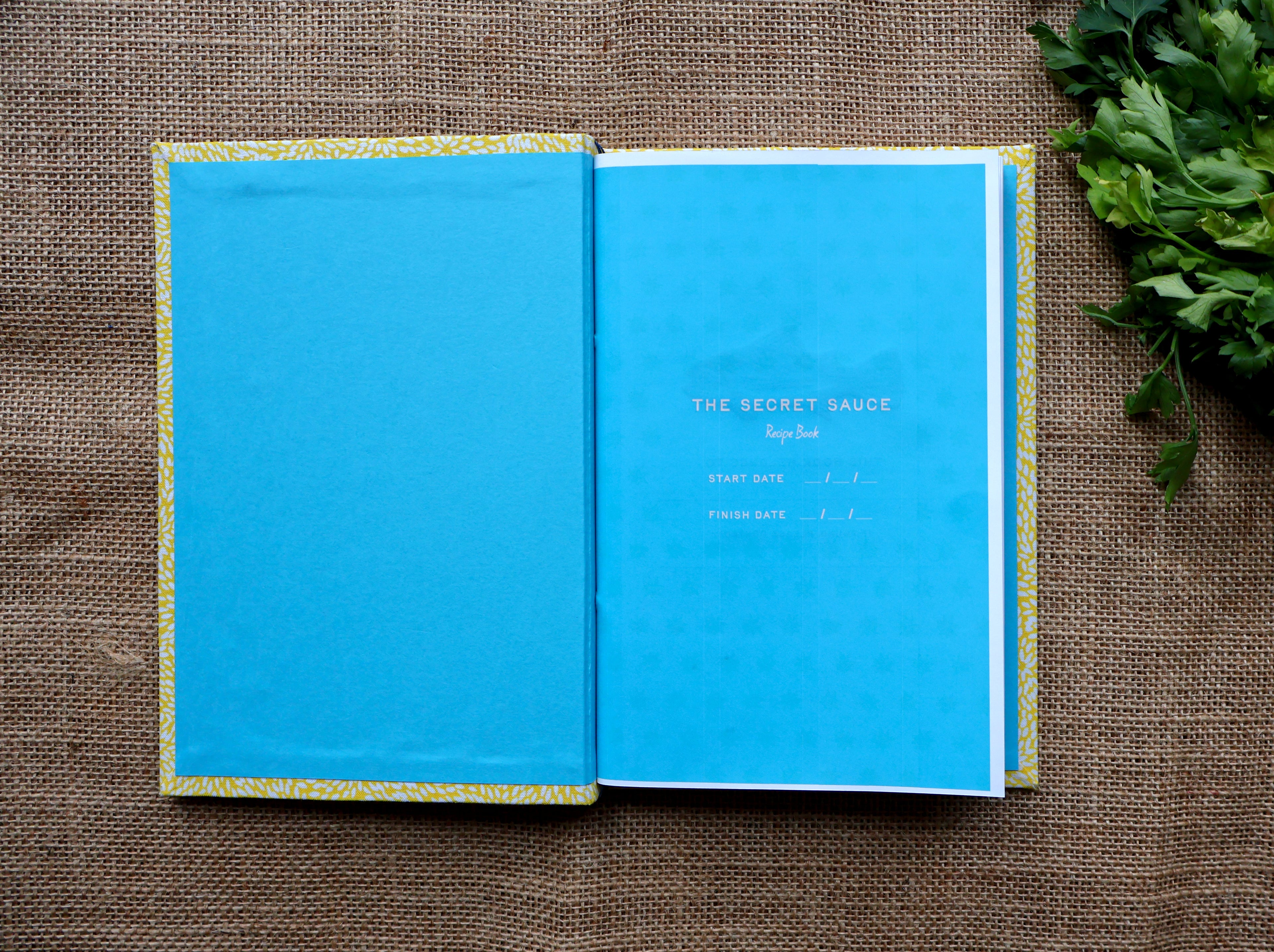 An open recipe log with bright blue pages, placed on a brown burlap surface with green leaves at the top right. The left page is solid blue, and the right page has small white text reading 'THE SECRET SAUCE recipe book.