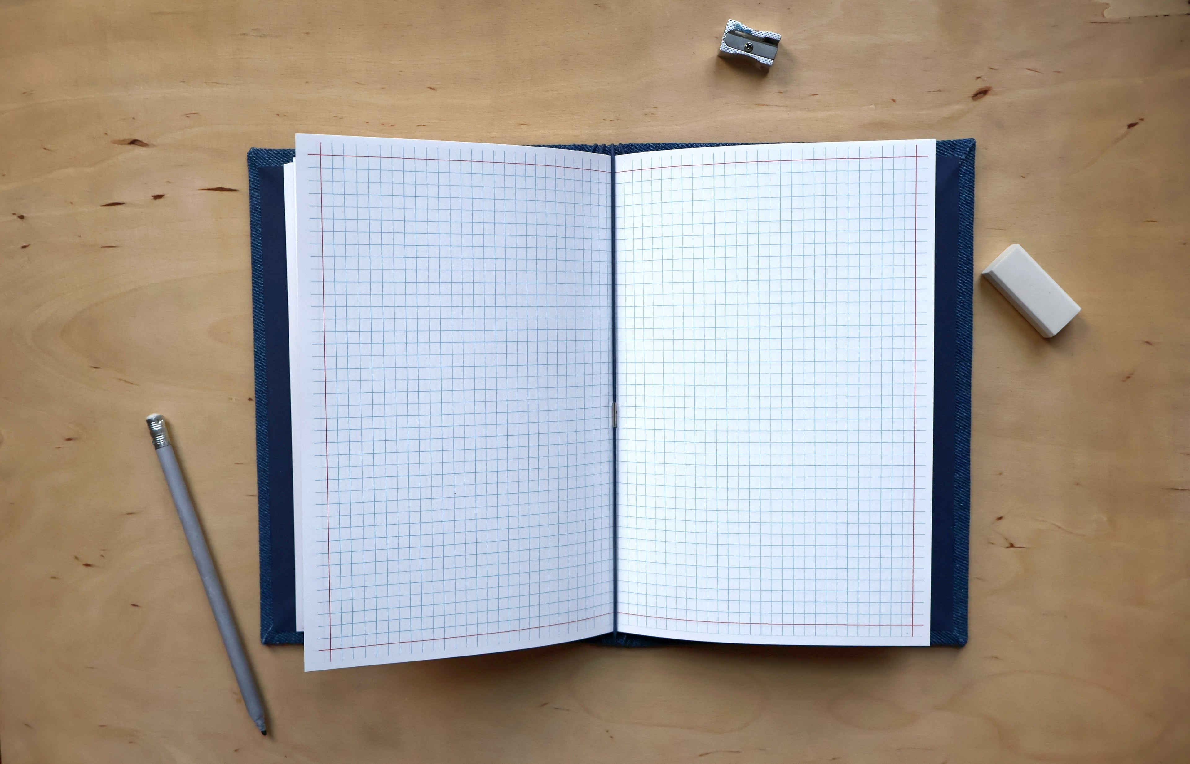 An open notebook with white graph paper pages and a dark navy blue cover, laid flat on a light wood desk with a pencil, eraser, and binder clip nearby.
