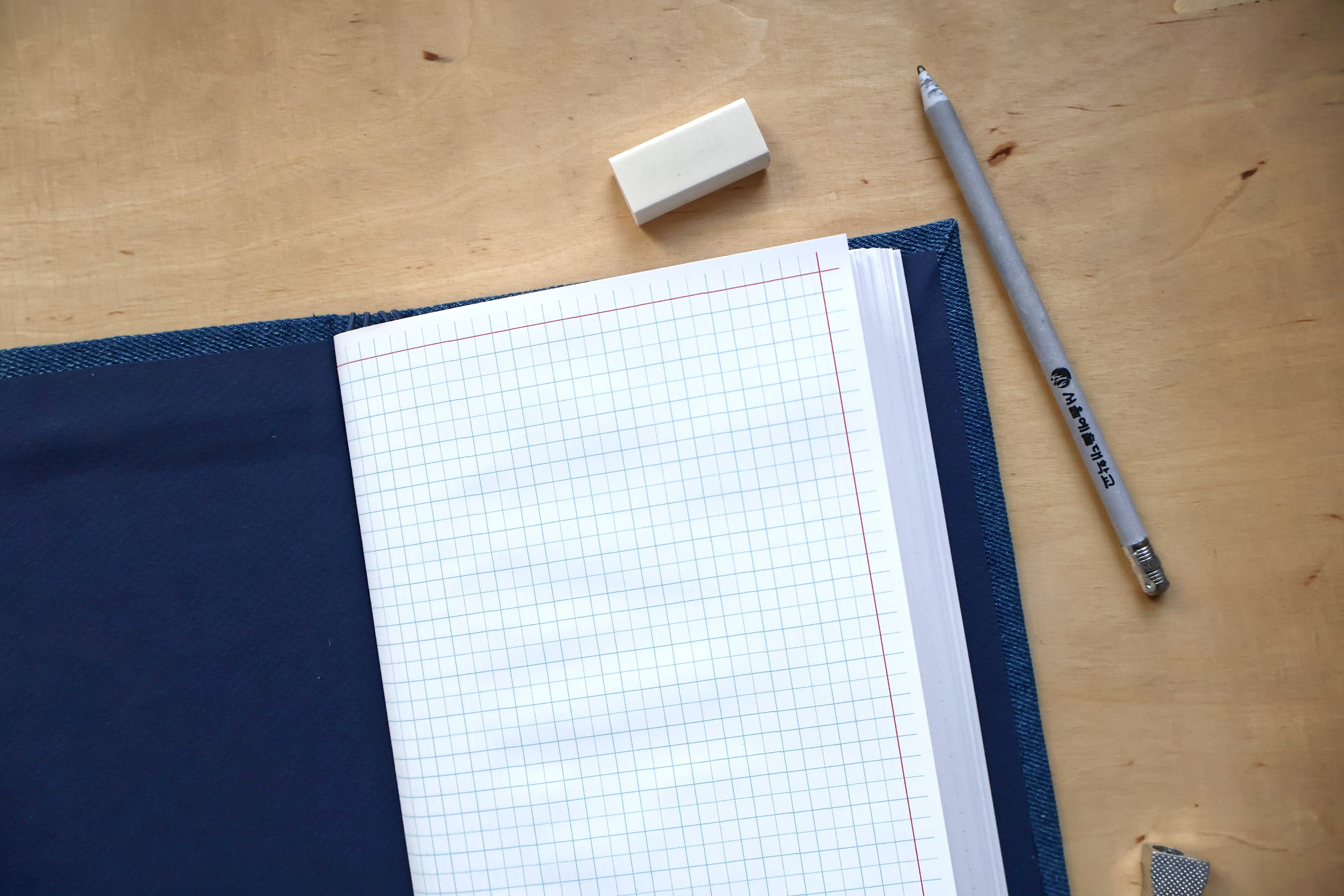 A top-down view of a navy blue notebook or planner cover with white graph paper pages, placed on a light wood desk alongside a gray Staedtler mechanical pencil, a small white eraser, and a small gray paper clip.