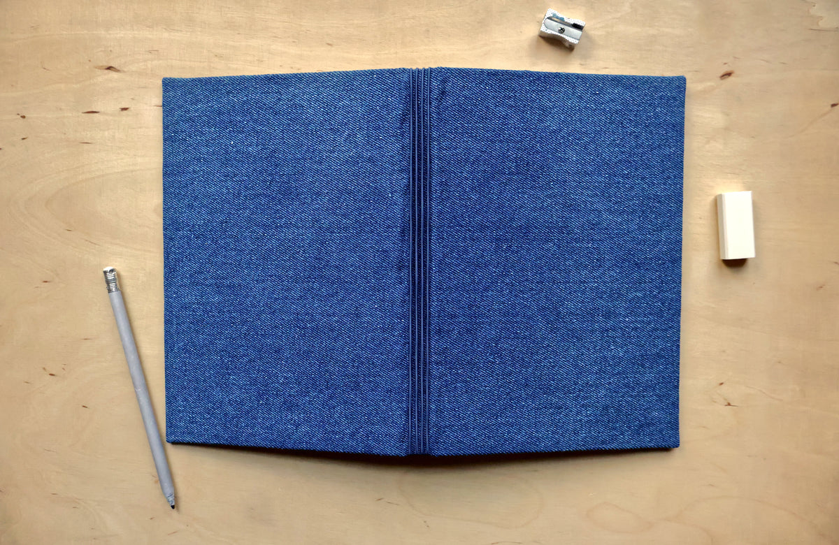 A blue fabric-covered notebook laid open on a light wood tabletop, with a pencil on the left and small stationery items on the right.