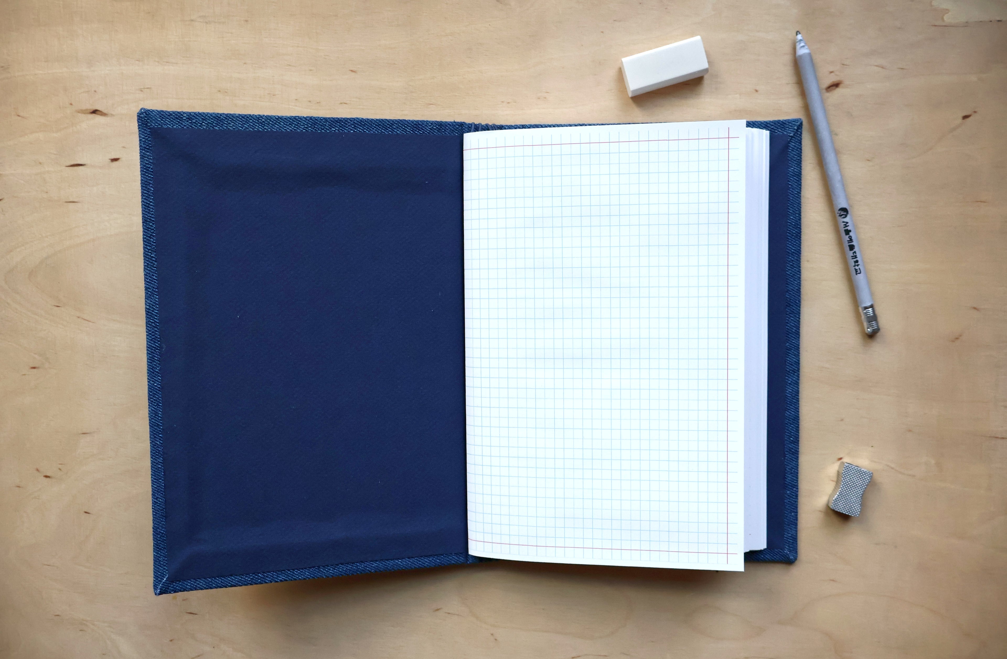 A navy blue denim-covered notebook with white graph paper pages, shown open on a light wood tabletop with a pencil, eraser, and small gray eraser piece nearby.