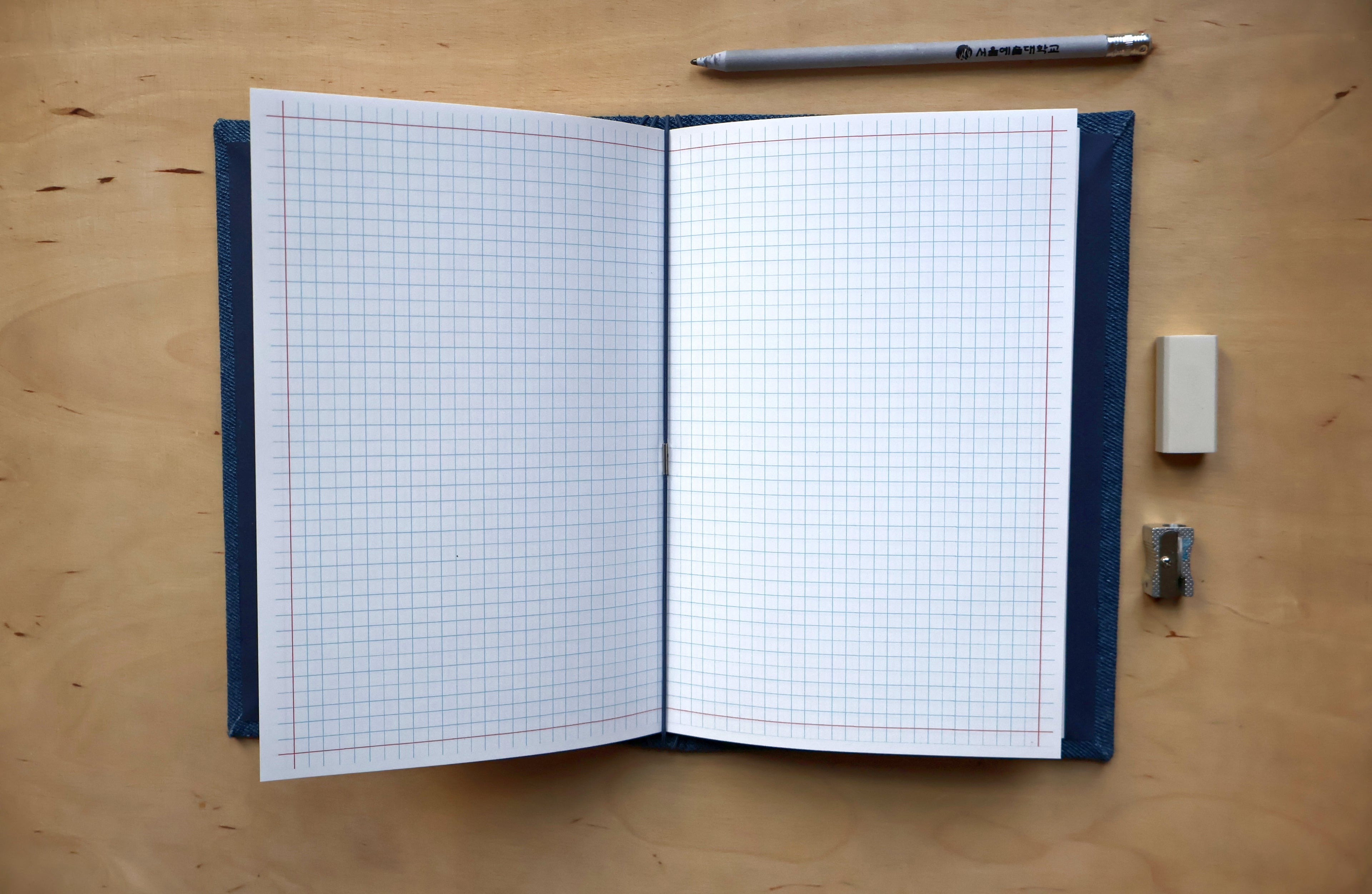 An open graph notebook with white grid pages and a dark blue cover, placed on a light wood desk with a pen, eraser, and pencil clip nearby.