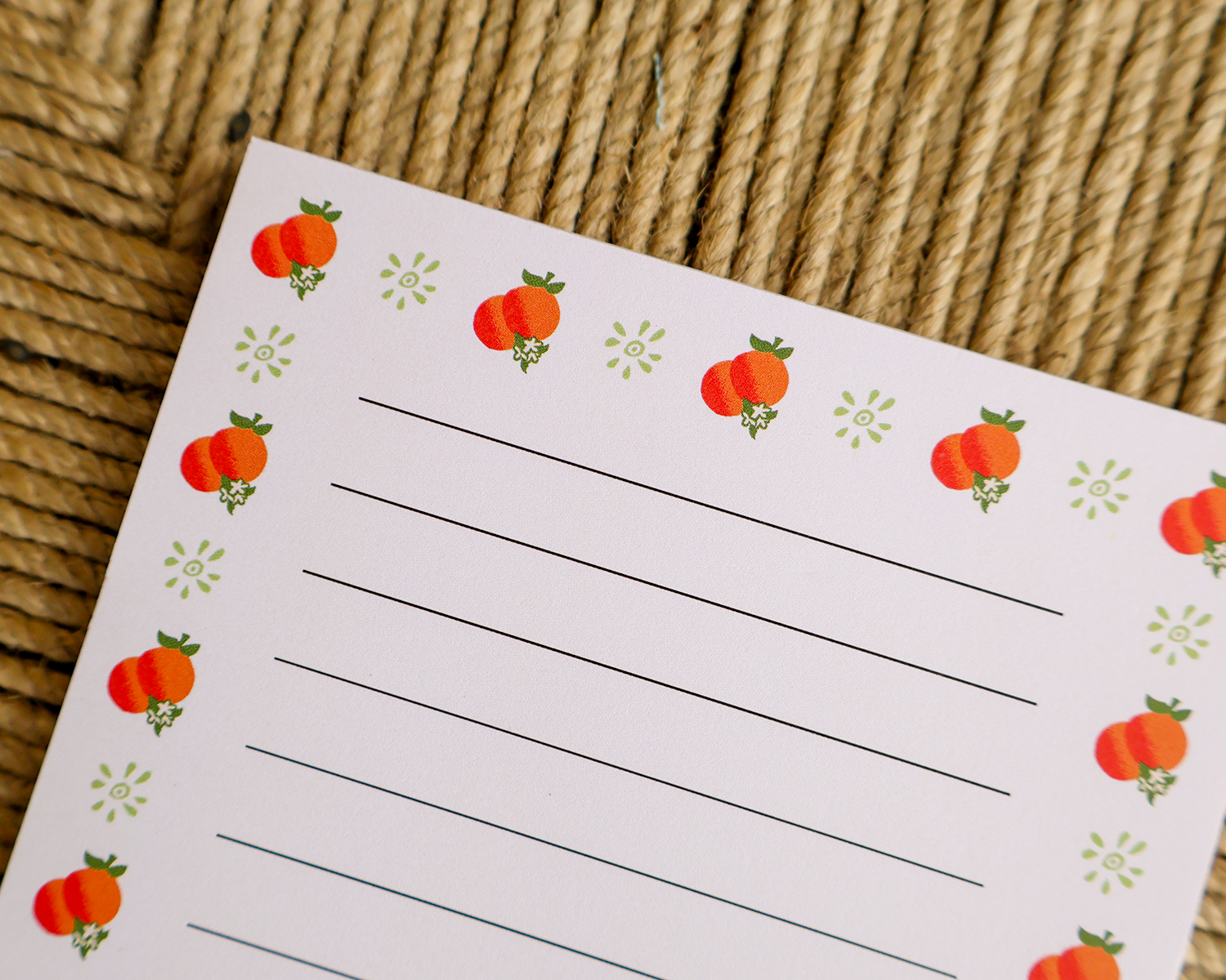 A close-up of a lined writing notepad with a white background, decorated with orange illustrations and small green floral motifs along the border, placed on a woven straw surface.