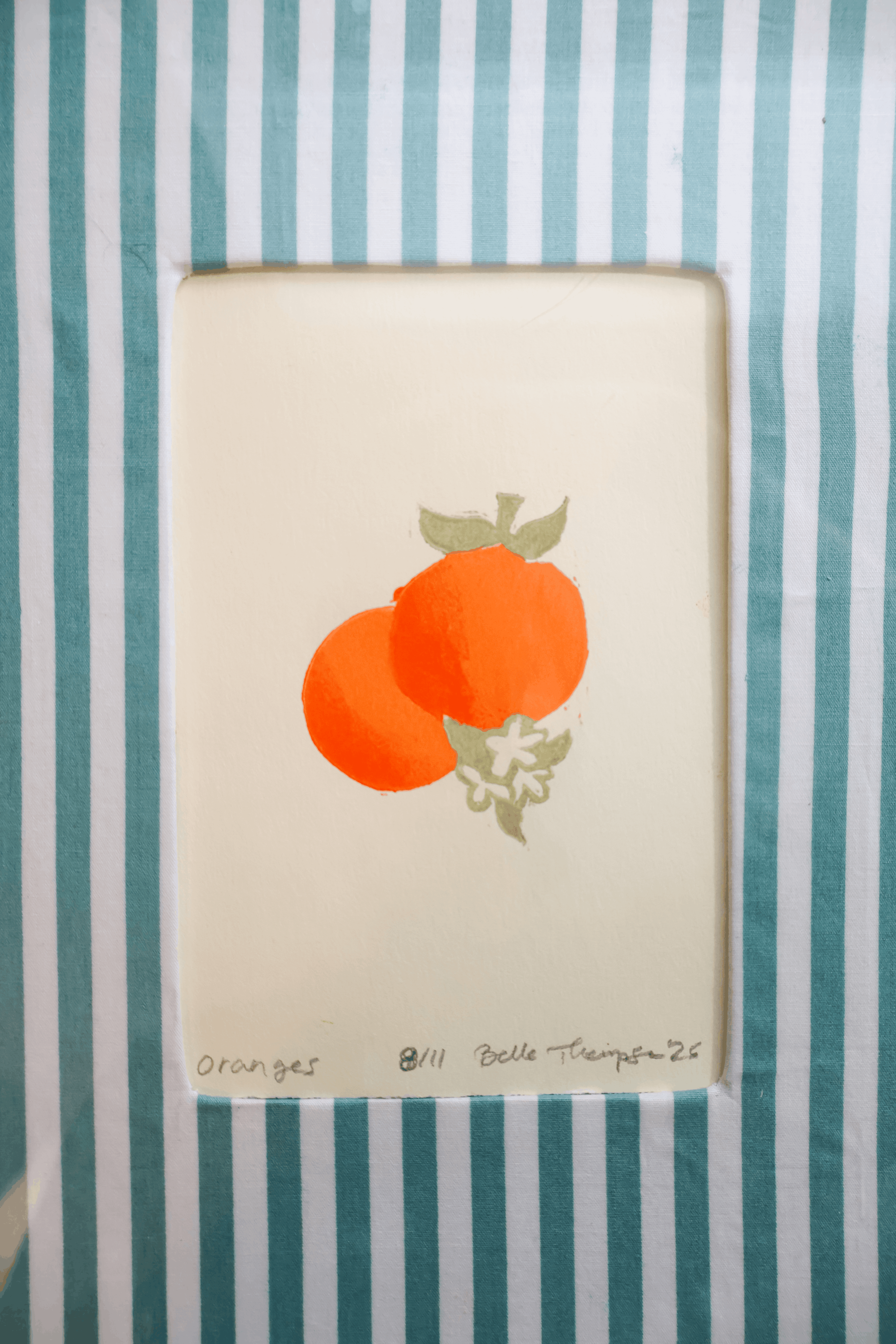 A square art print featuring two orange fruits with green leaves on an off-white background, placed on a teal and white striped surface. Handwritten text at the bottom reads 'ORANGES' and 'Belle Thompson 25'.