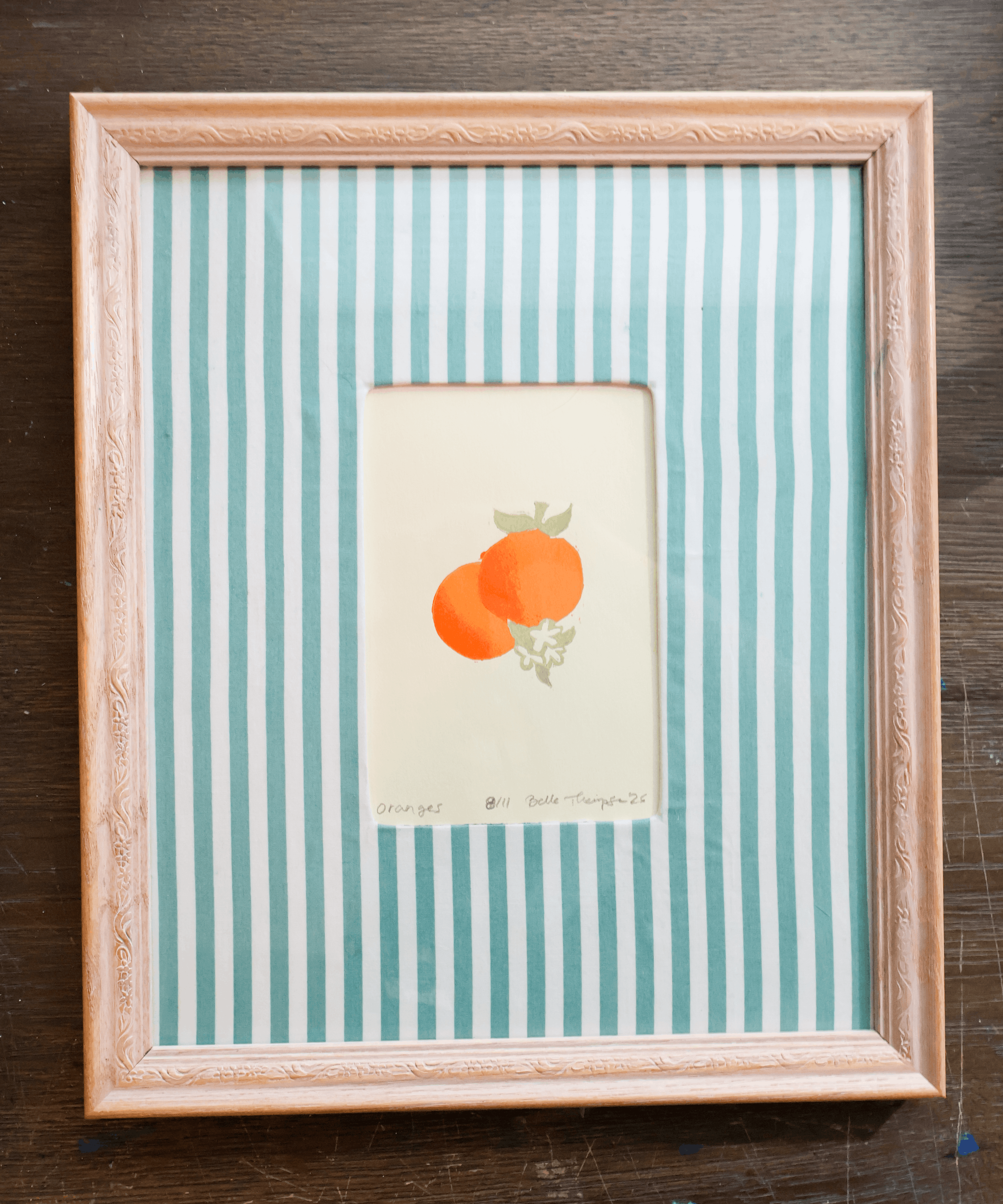 A framed art print featuring two orange fruits on a cream background, set against a mint green and white striped mat and enclosed in a light wood frame, placed on a dark wooden surface.