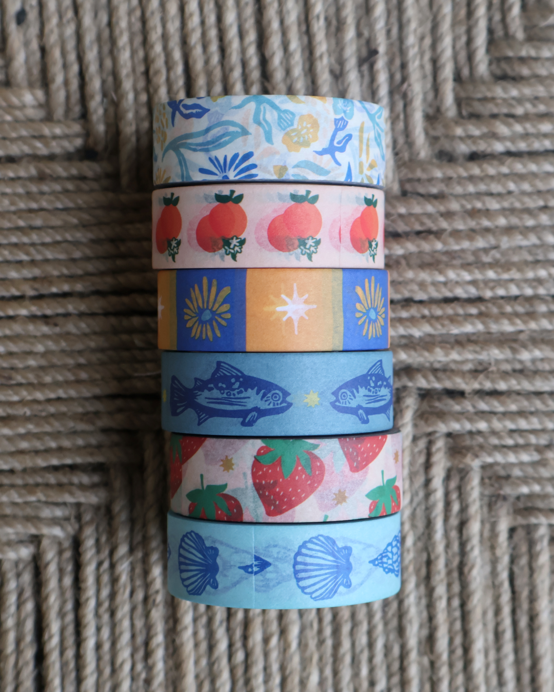 linocut design of linocut floral, orange citrus, checkered floral, trout blue fish, red strawberries, and blue seashells on decorative washi tape. Showing the design on a textured brown background. by dovetail studio
