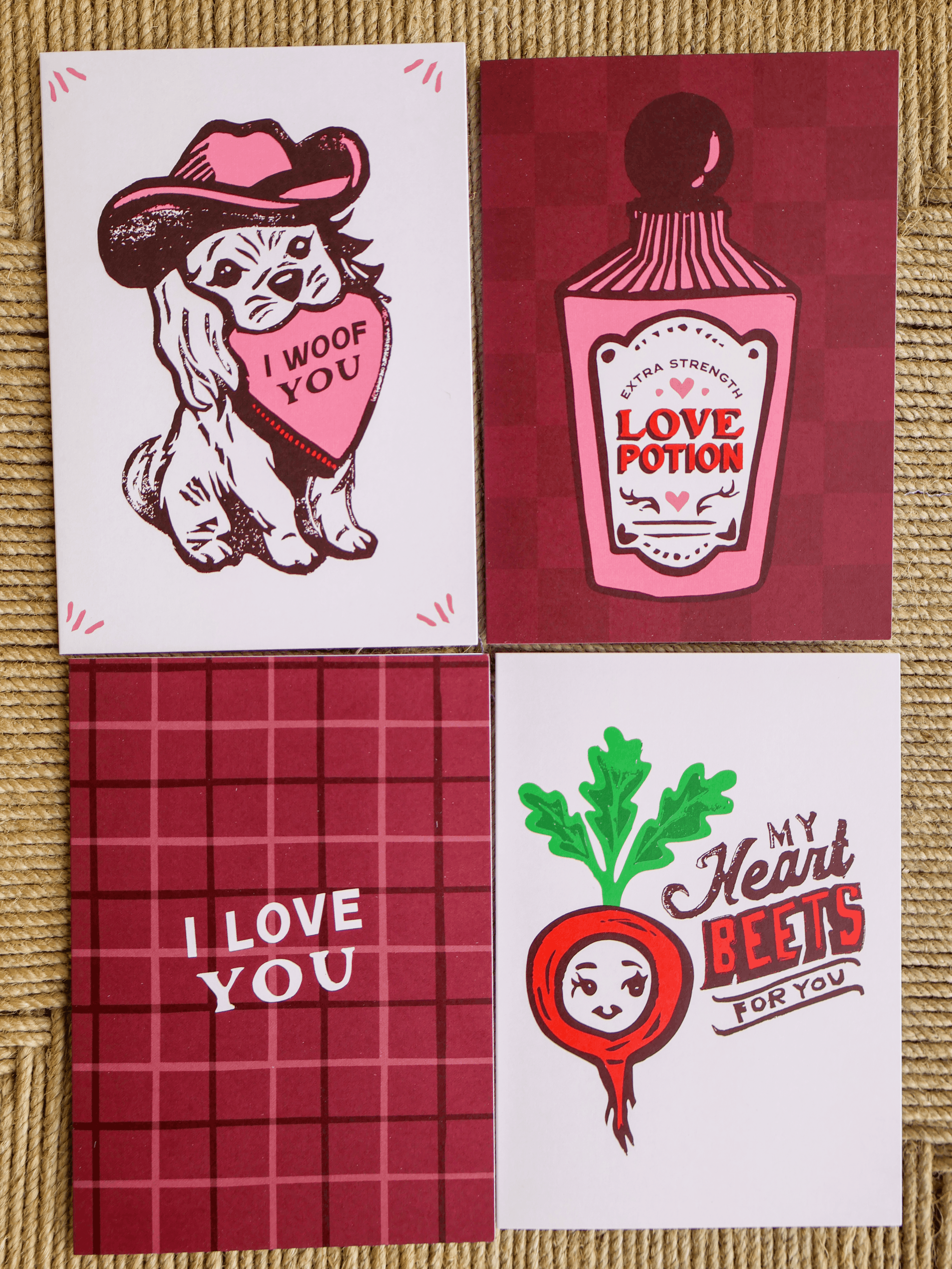 Four square greeting cards arranged in a 2x2 grid on a woven tan surface. The cards feature playful Valentine-themed illustrations and phrases: a white card with a dog wearing a hat and heart that says 'I WOOF YOU'; a dark red card with a pink bottle labeled 'LOVE POTION'; a dark red card with 'I LOVE YOU' in white text; and a white card with a red beet illustration and the text 'My Heart BEETS FOR YOU'.