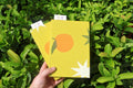A hand holds two yellow textured notebooks or booklets decorated with orange and white starburst illustrations, set against a background of green leaves. Hand-printed linocut cover, hand bound self care reflections log