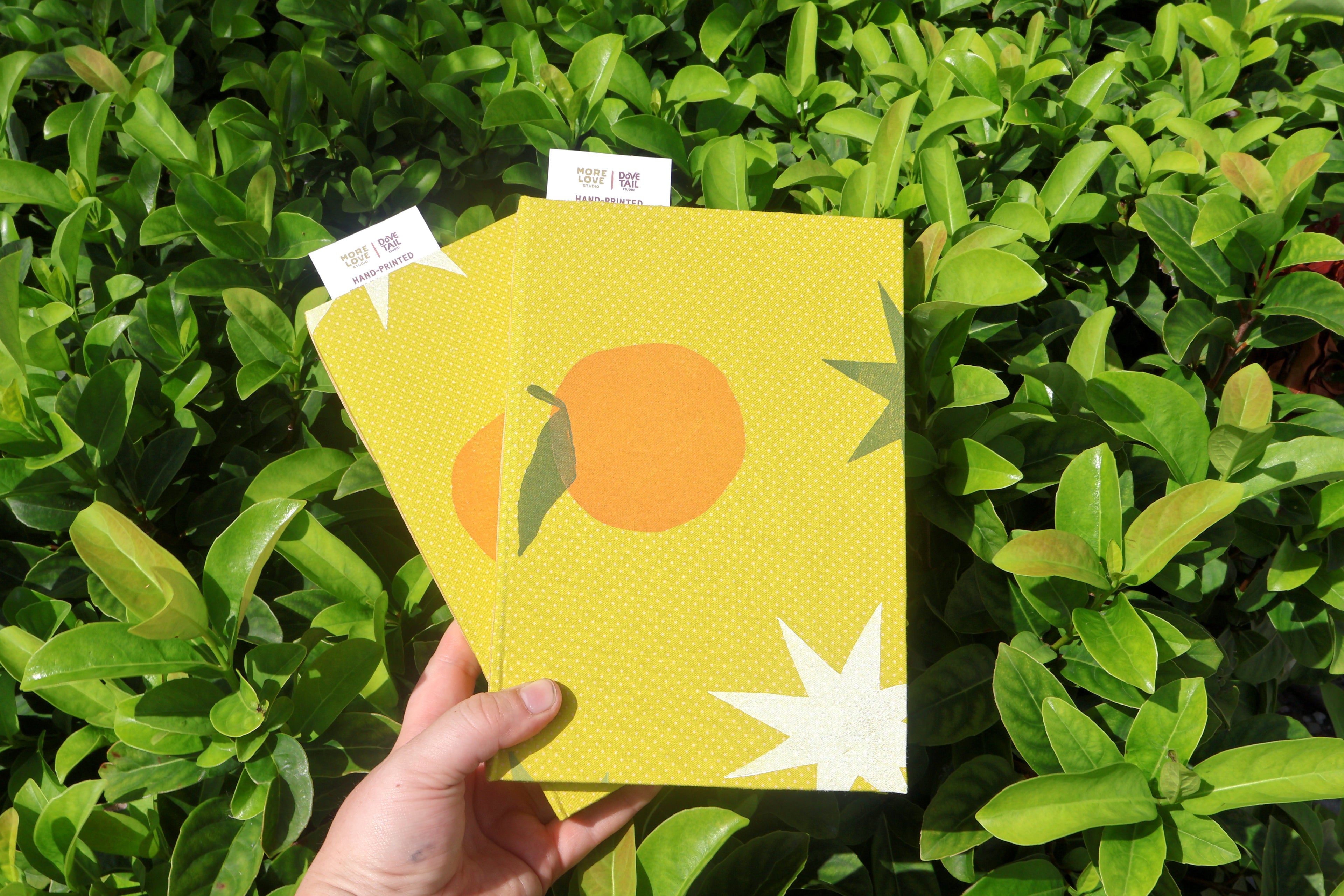 A hand holds two yellow textured notebooks or booklets decorated with orange and white starburst illustrations, set against a background of green leaves. Hand-printed linocut cover, hand bound self care reflections log