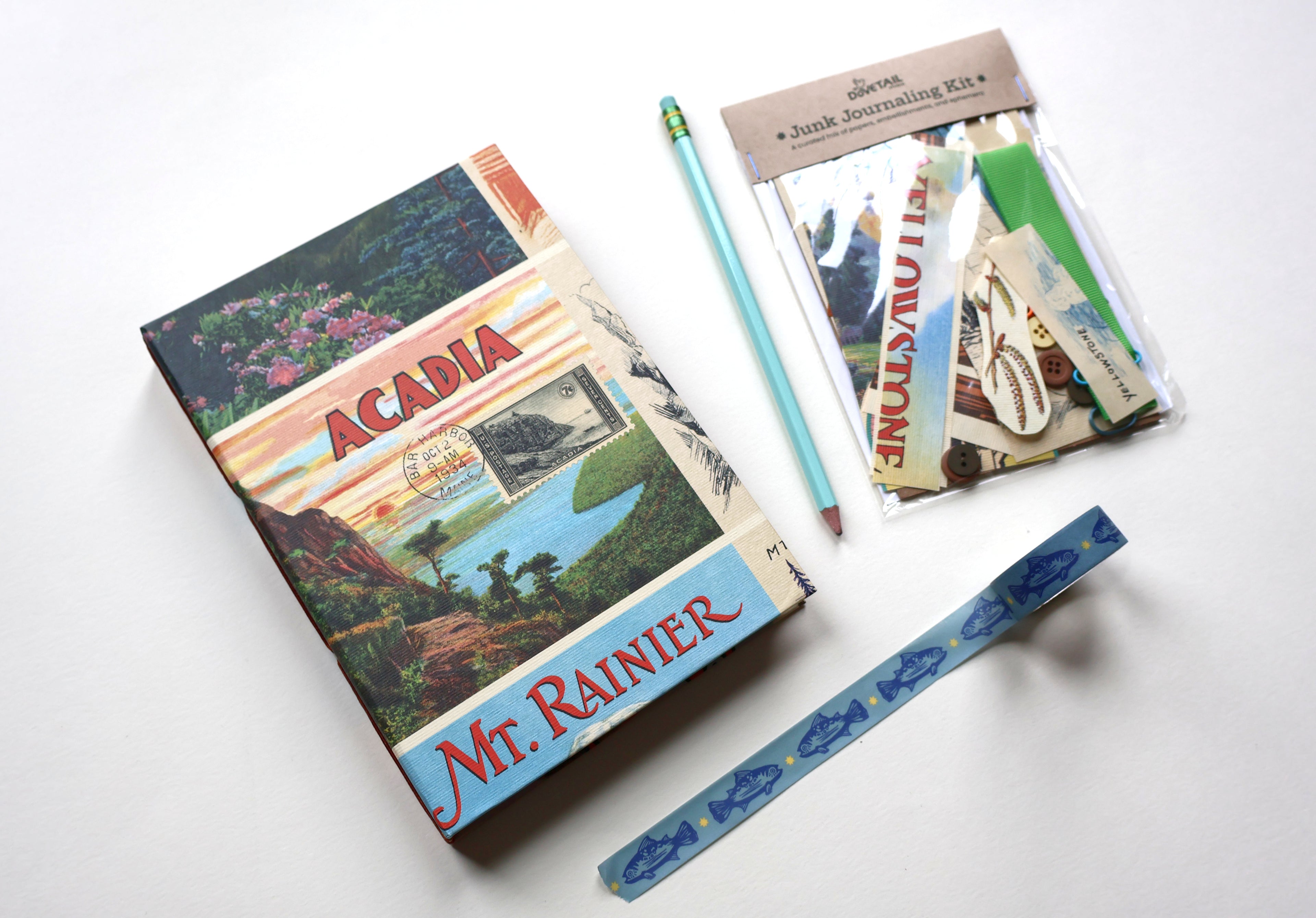 A journaling kit featuring a vintage-style 'Acadia Mr. Rainier' sketchbook, a light blue pencil, and a clear plastic bag of scrapbook/journaling items, and blue trout patterned washi tape arranged on a white background.