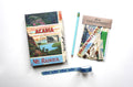 A flat-lay of a junk journaling kit on a white background, including an 'Acadia Mt. Rainier' sketchbook, a light blue pencil, a bag of assorted paper scraps, and a roll of blue trout-patterned washi tape.