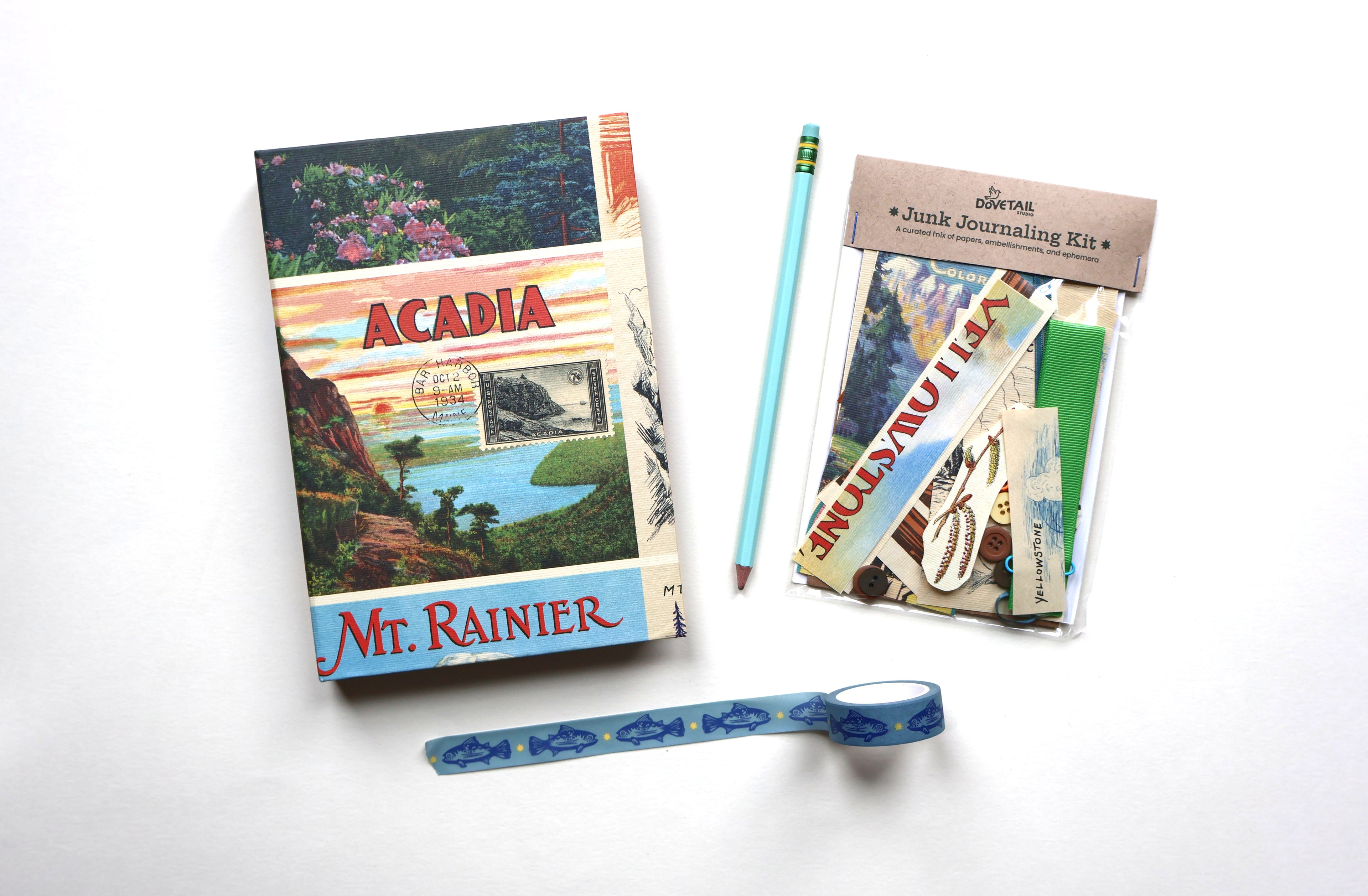 A flat-lay of a junk journaling kit on a white background, including an 'Acadia Mt. Rainier' sketchbook, a light blue pencil, a bag of assorted paper scraps, and a roll of blue trout-patterned washi tape.