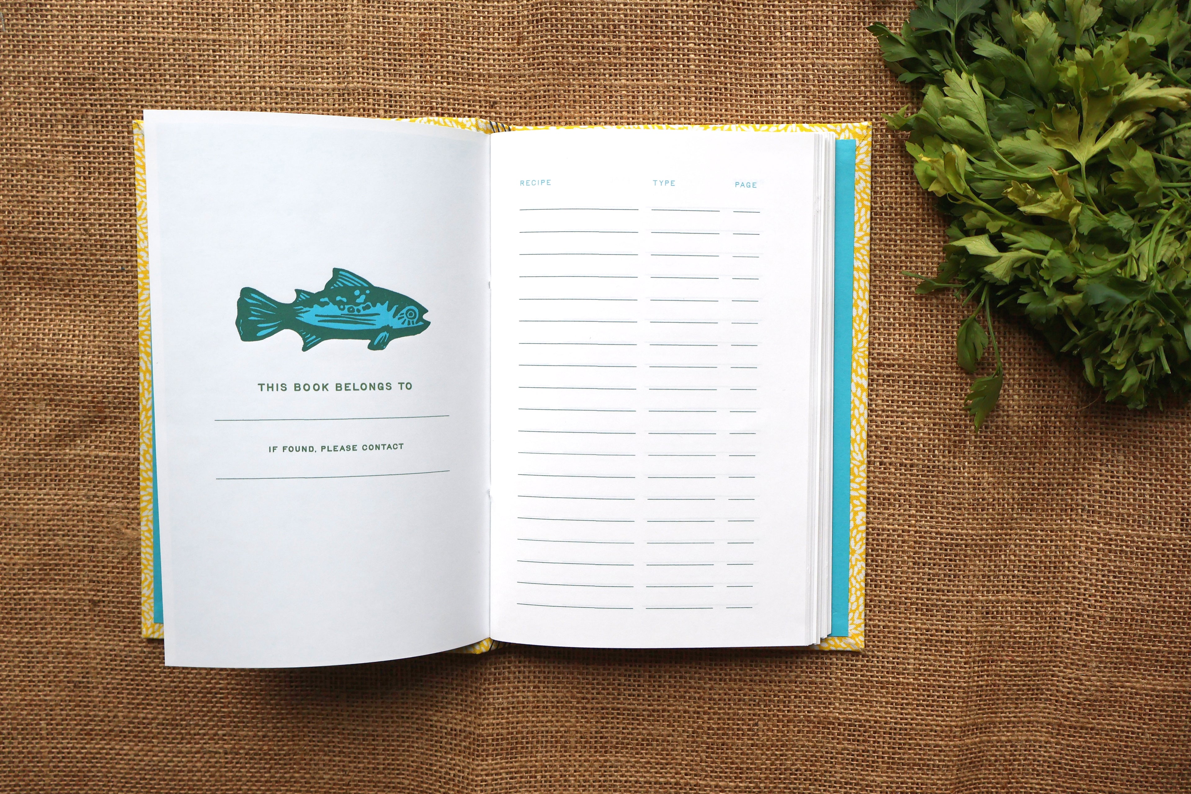 An open hardcover recipe log lies on a brown burlap surface, with a teal fish illustration on the left page and lined interior pages on the right. A bunch of green herbs sits at the upper right.