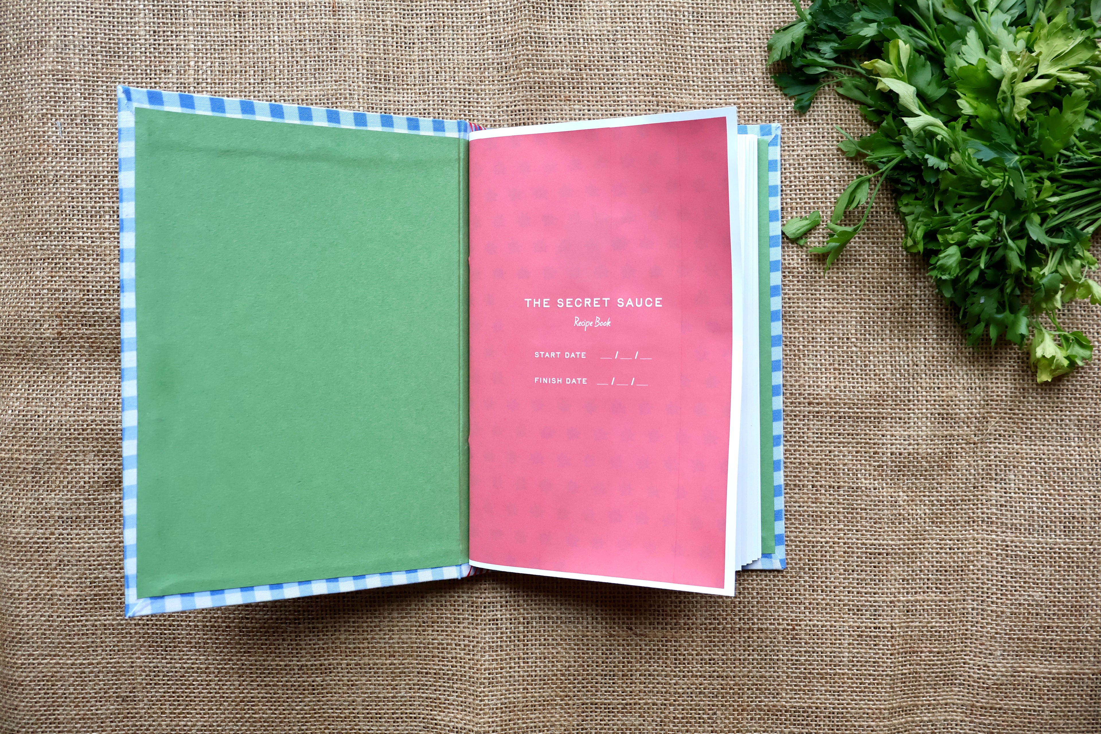 An open recipe book with a green left page, a pink right page titled 'THE SECRET SAUCE Recipe,' and a blue-and-white checkered cover edge, photographed on burlap with green herbs in the upper right.
