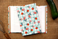 A fabric recipe log with a peach and gingham pattern, shown laid over an open lined recipe log on a burlap surface, with a cucumber and whisk as props. Handmade original design