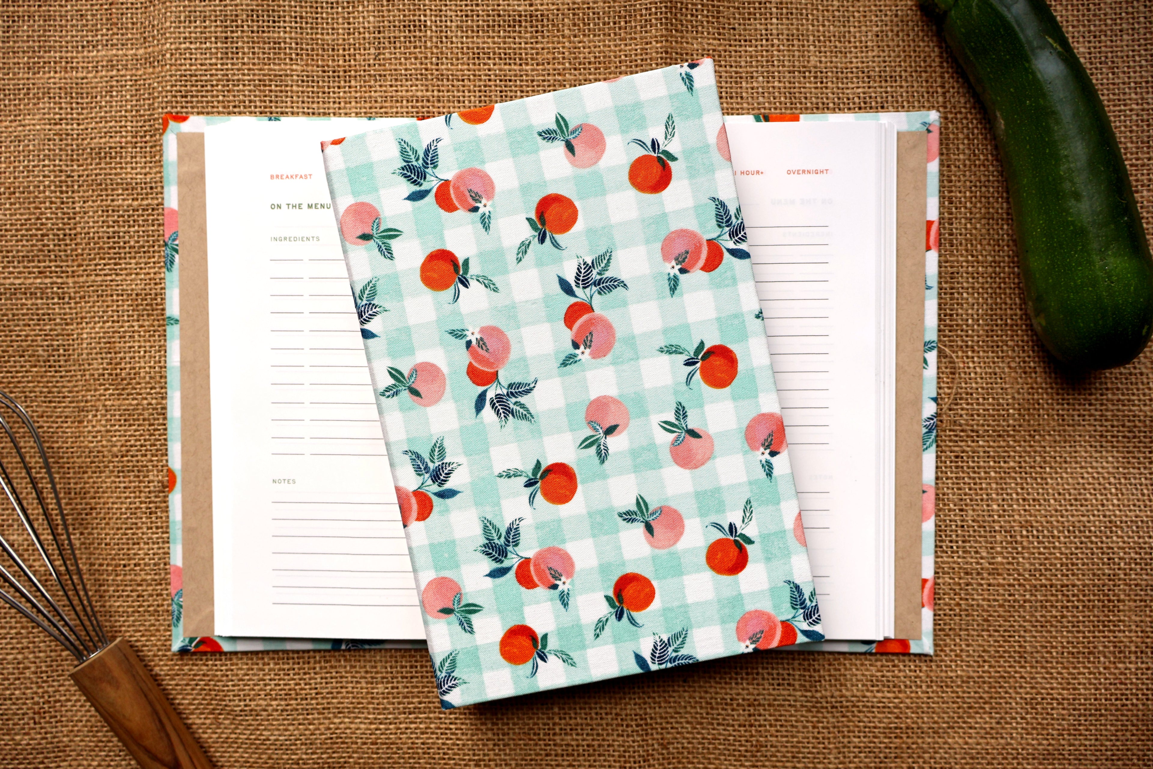 A fabric recipe log with a peach and gingham pattern, shown laid over an open lined recipe log on a burlap surface, with a cucumber and whisk as props. Handmade original design