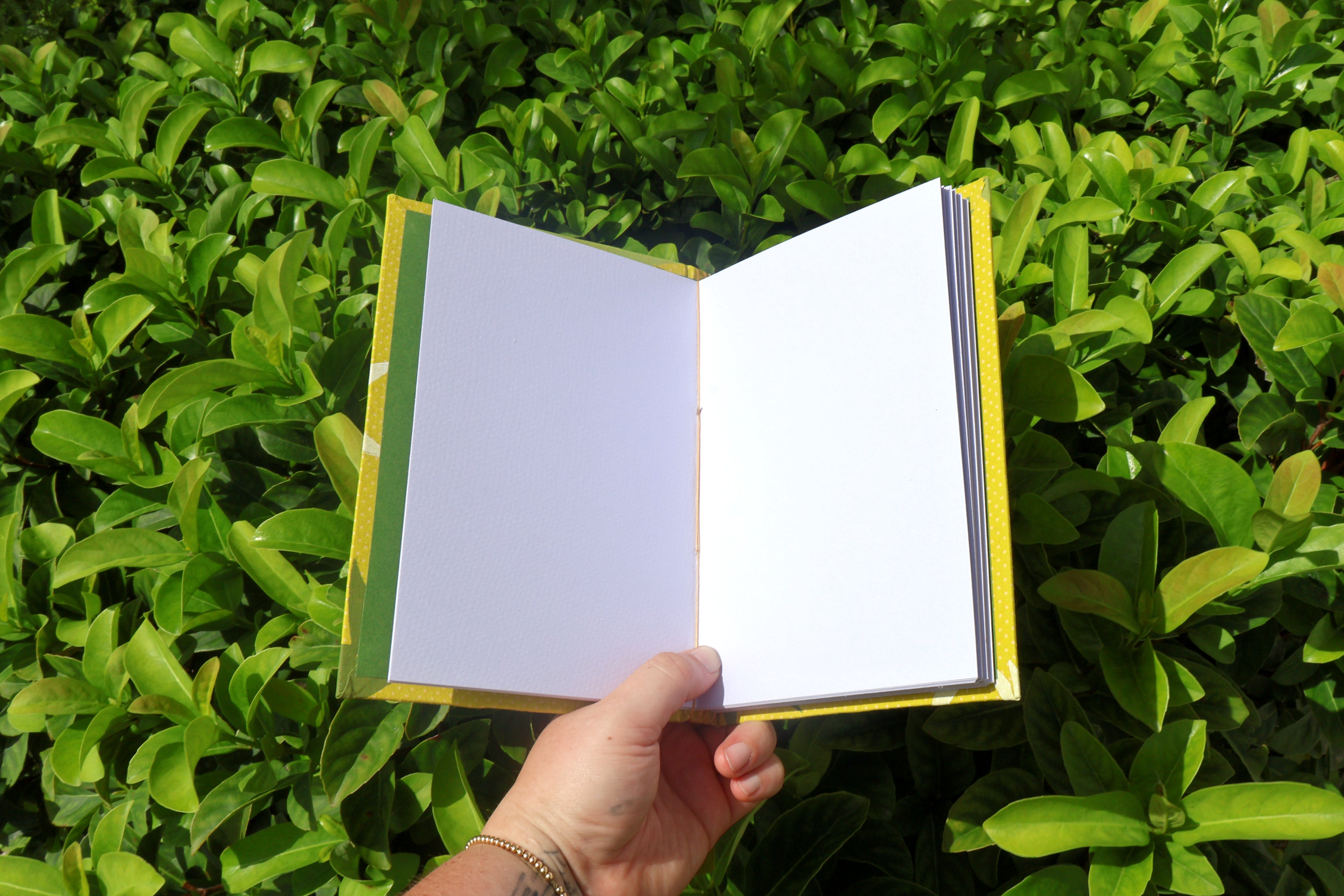 A hand holds an open, green, hand-bound and printed self-care reflection logs with white starburst shapes, photographed outdoors against dense green leaves. Hand bound and linocut printed with more love studio