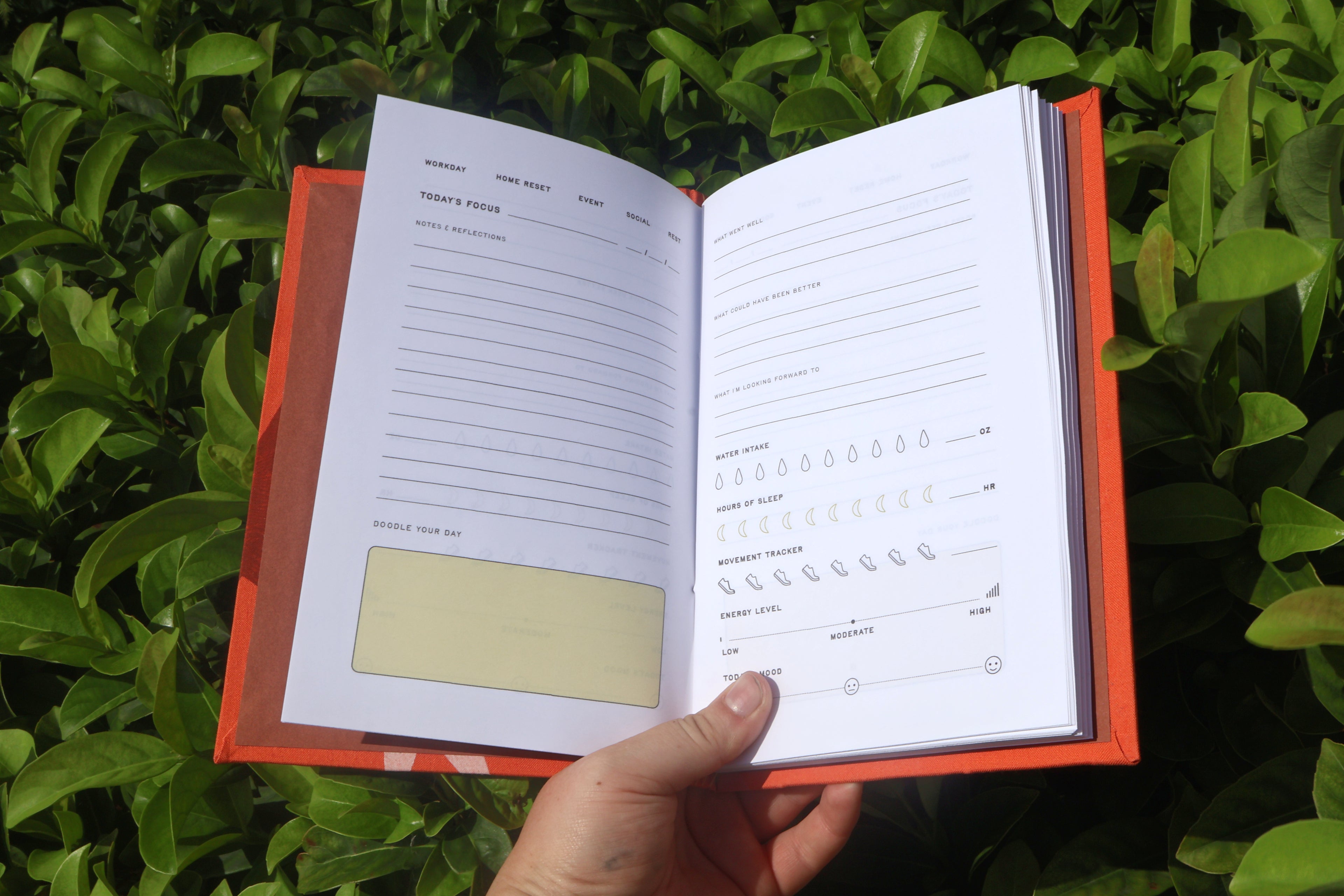An open habit tracker self care journal with white pages and a bright orange cover, held by a hand against a background of green foliage. Hand-bound and printed with more love studio
