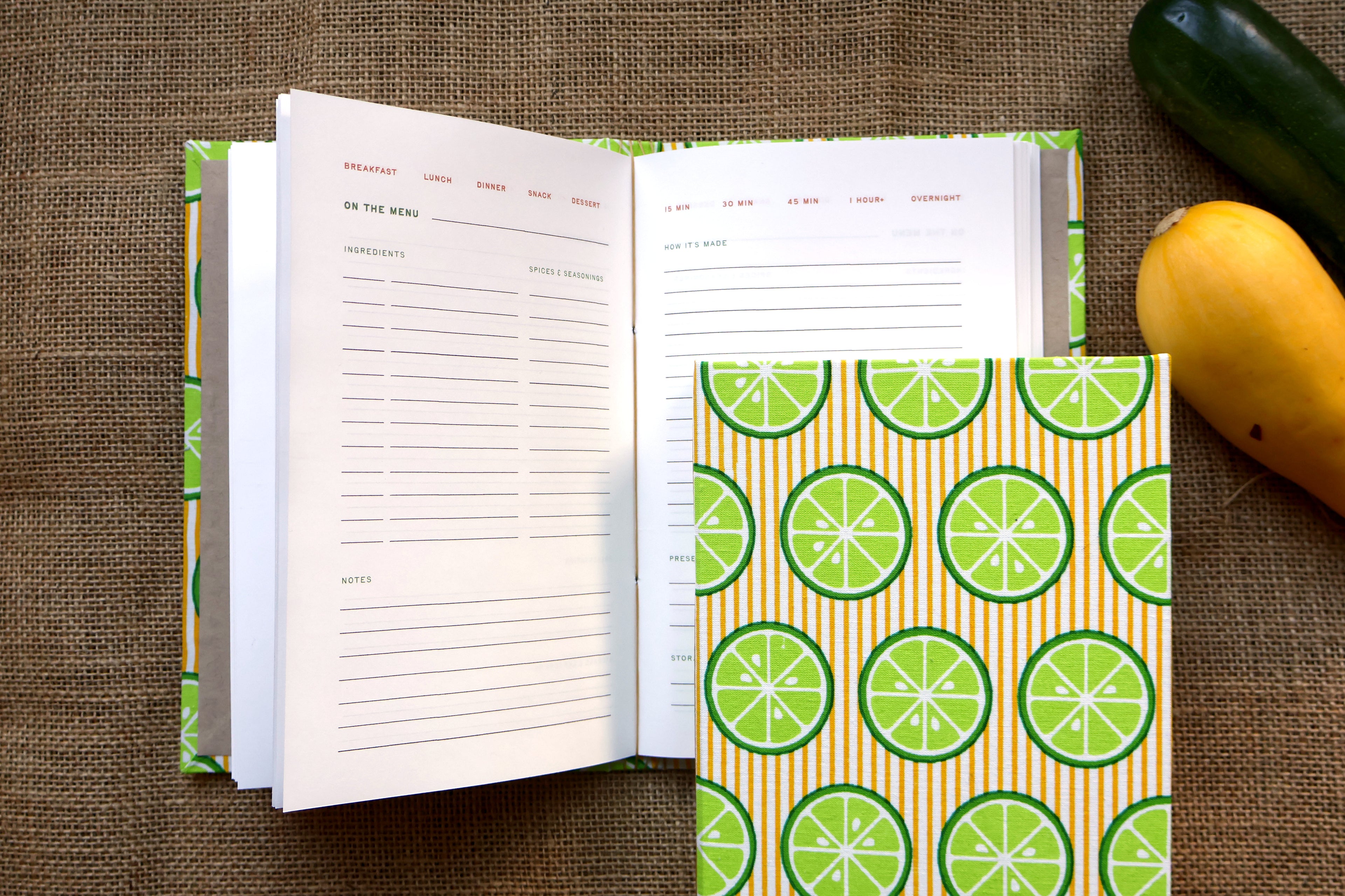 An open recipe log and matching hardcover notebook with a lime slice pattern, styled on a burlap surface with a cucumber and a yellow squash nearby.