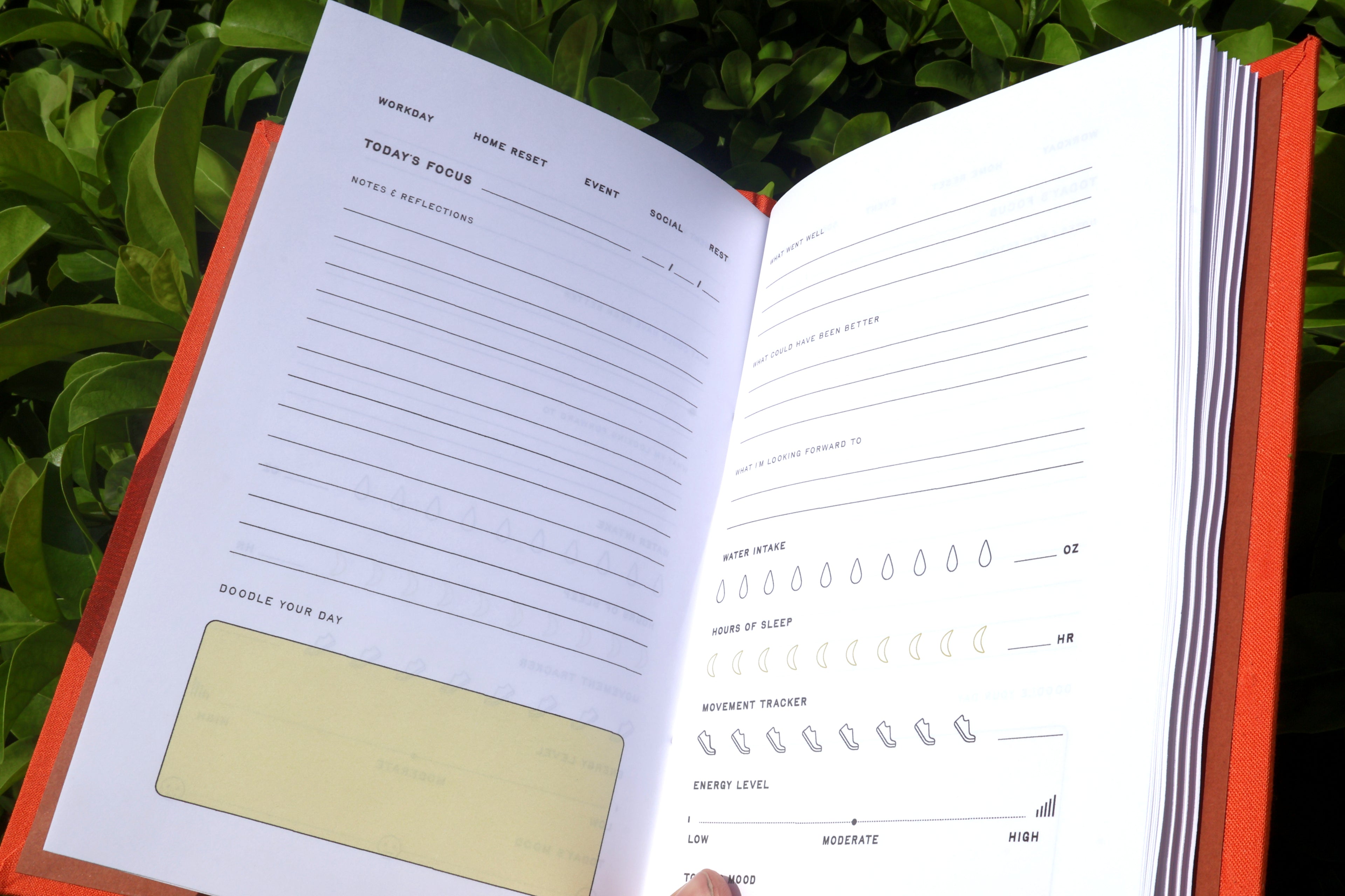 An open habit tracker self care reflections log journal with white pages showing daily reflection prompts and habit tracking lines, set against a background of green foliage. hand bound and linocut printed with more love studio