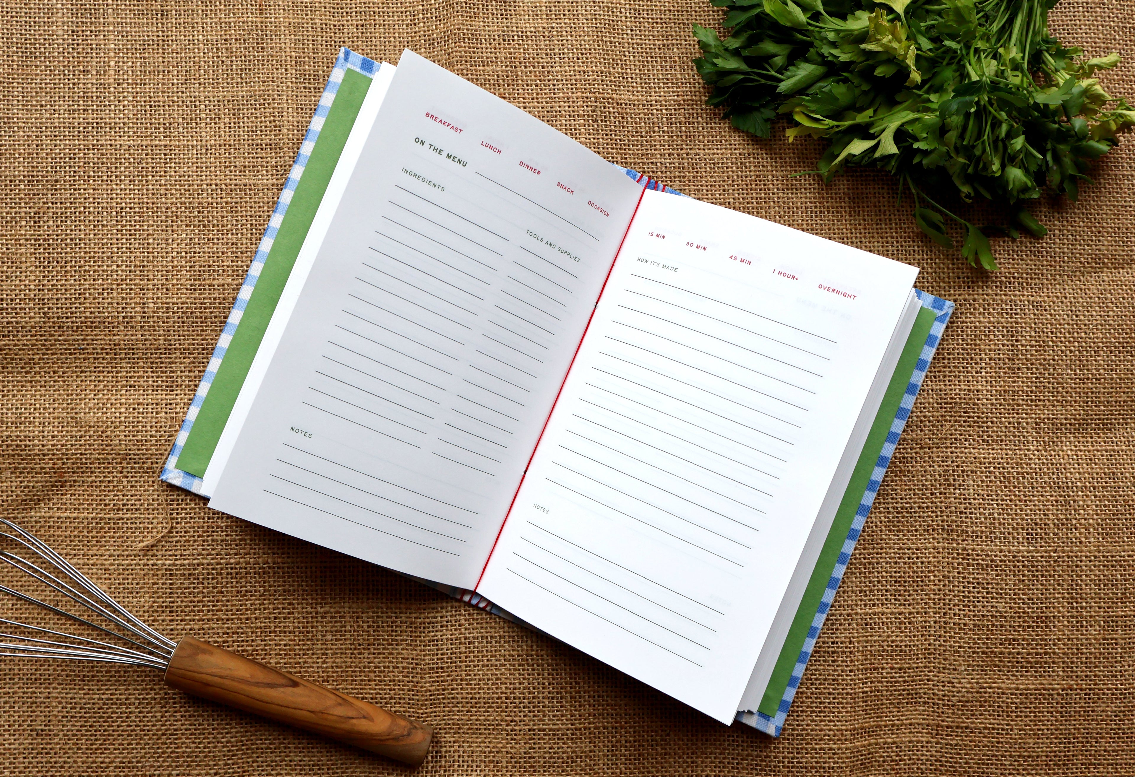 An open recipe journal with lined pages, blue and green checkered cover edges, and ared thread binding, styled on a burlap surface with a whisk, parsley, and a wooden spoon handle.