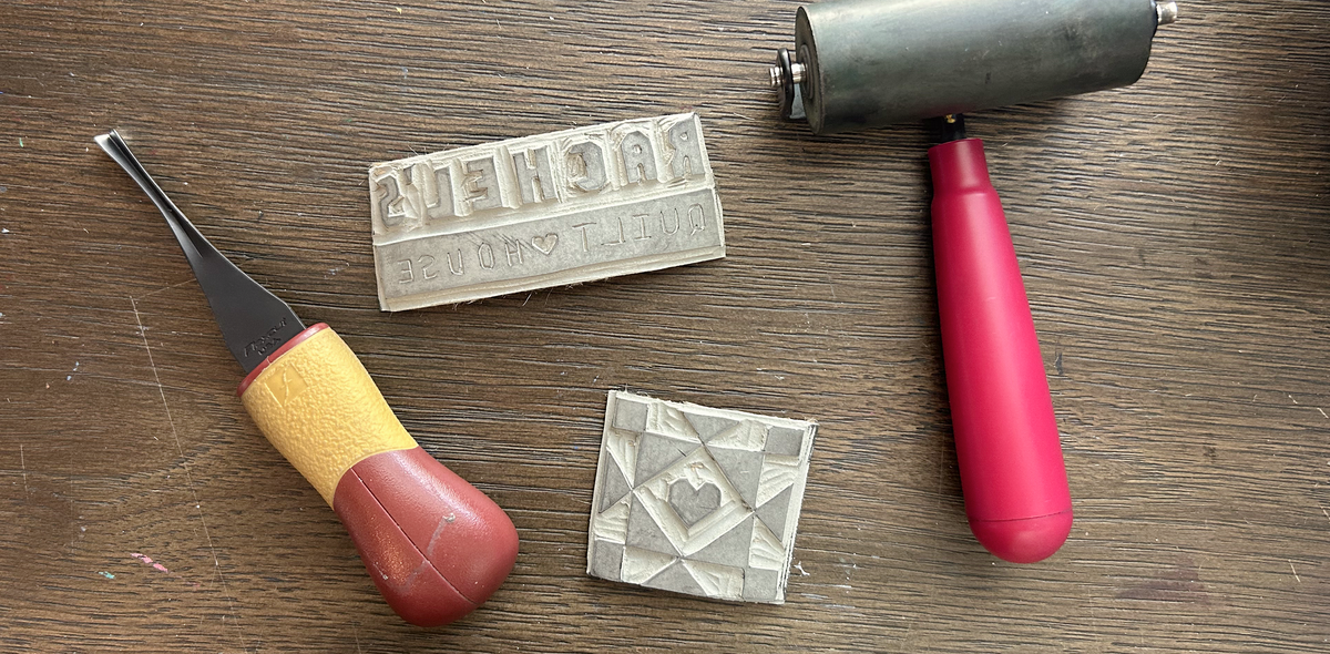 A flat-lay of four quilting tools on a wooden surface: a wooden text block stamped 'rachels quilt house', a wooden geometric block, a metal roller with a red handle, and a wooden chisel with a yellow and red handle. Showing the hand carved linocut master copy of the custom logo