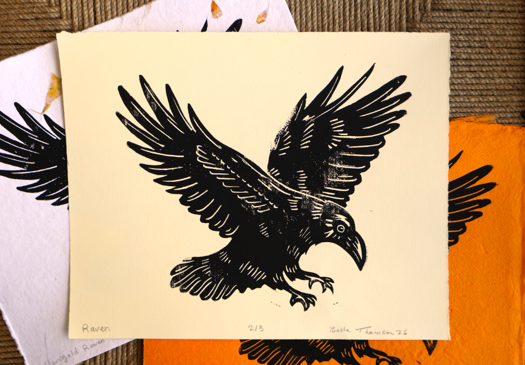 A black ink bird print on cream paper, shown on top of other art prints with orange and white paper visible underneath.