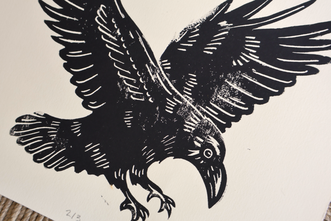 A black and white woodcut-style print of a raven in flight, shown close up on a white paper background with a small portion of a textured surface visible at the bottom left.