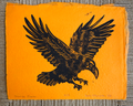 An orange art print or card featuring a black bird of prey illustration, photographed flat against a gray textured fabric background.