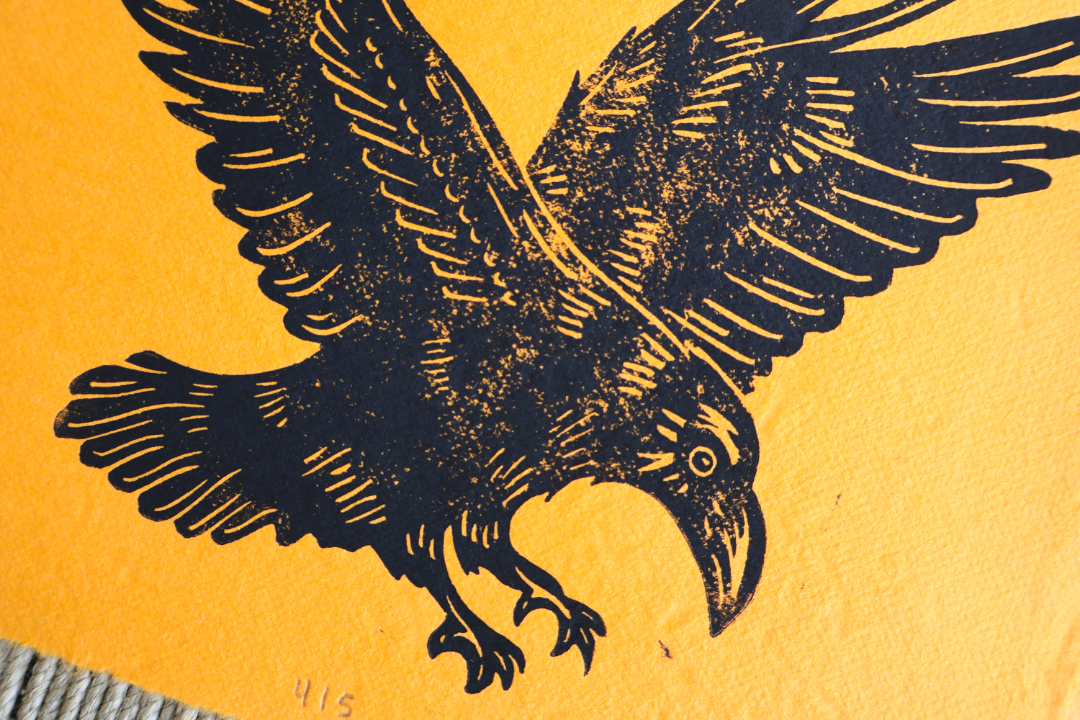 A close-up of a textured yellow-orange surface featuring a black printed illustration of a raven or crow with wings spread, plus a small gray textured strip at the lower left corner.