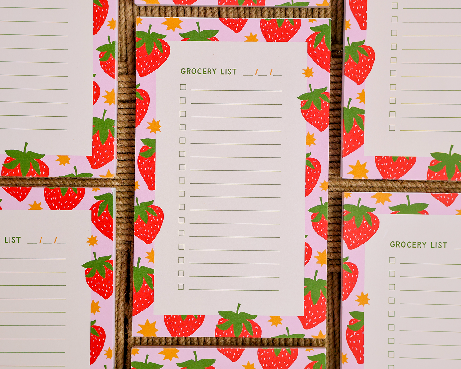 A close-up of a printed grocery list pad with a pink strawberry border, laid over a woven brown basket or mat. Multiple copies of the pad are visible in a repeating pattern. Linocut Design