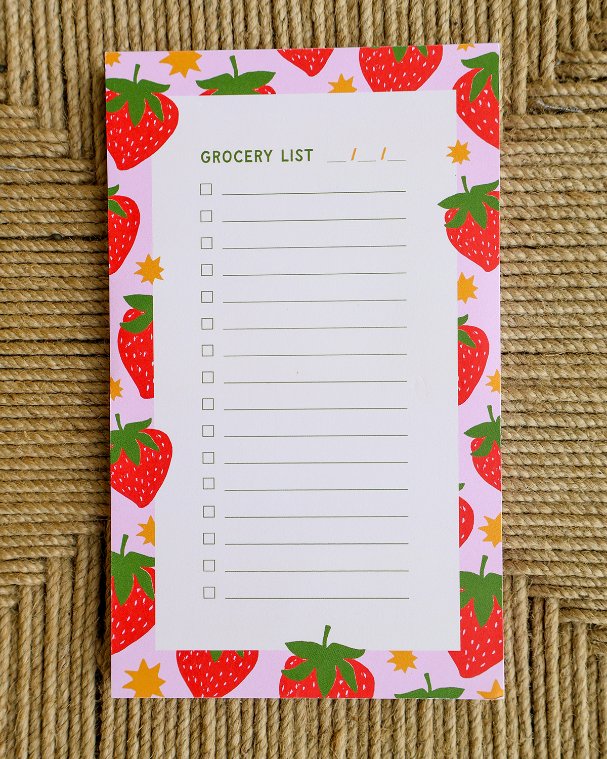 A rectangular notepad with a white center labeled 'GROCERY LIST' and lined checkboxes, surrounded by a pastel pink border printed with red strawberries, green leaves, and small yellow stars, placed on a woven tan surface.