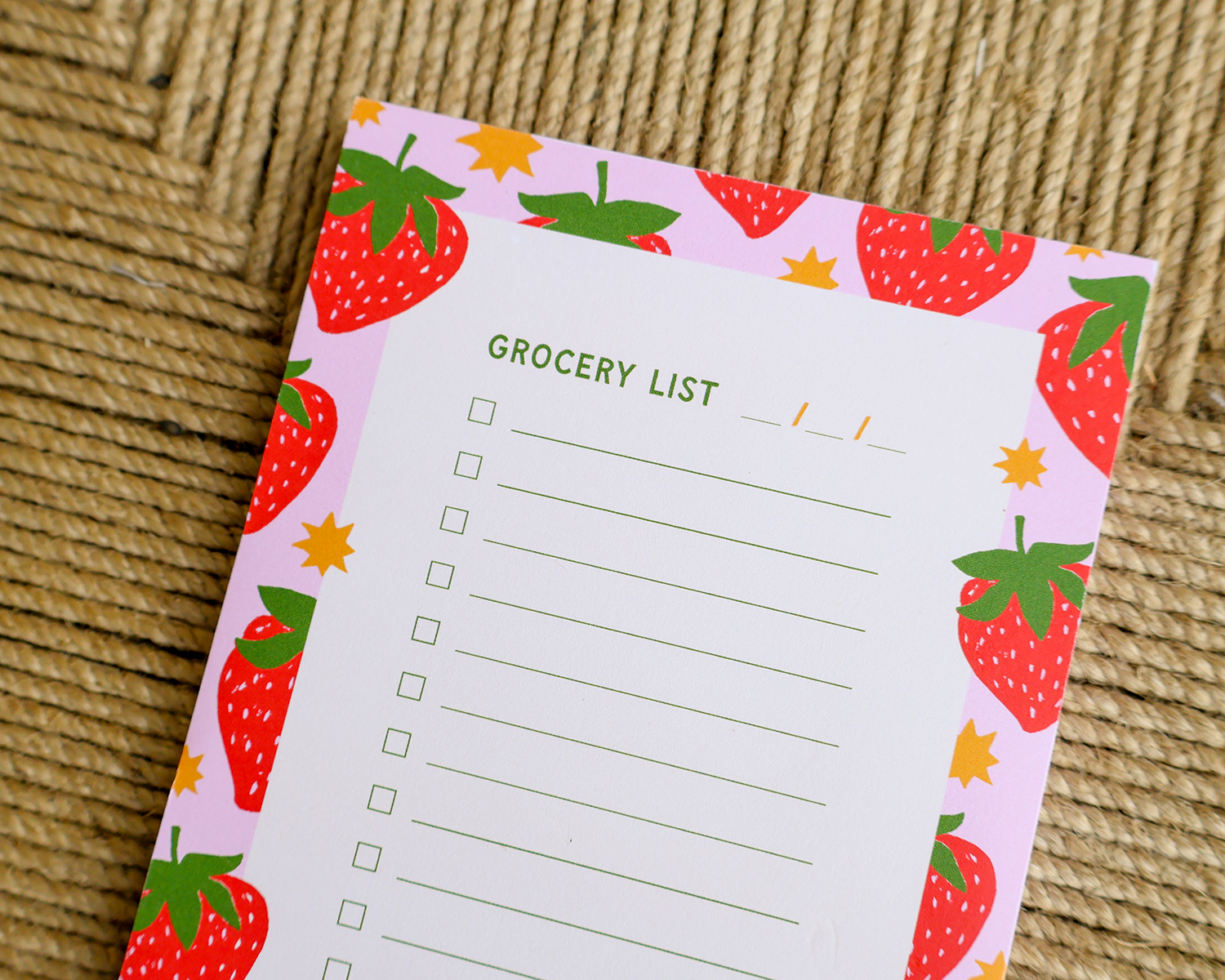 A strawberry-themed grocery list notepad with a pink border, red strawberries, green leaves, and yellow star accents, placed on a woven straw surface.