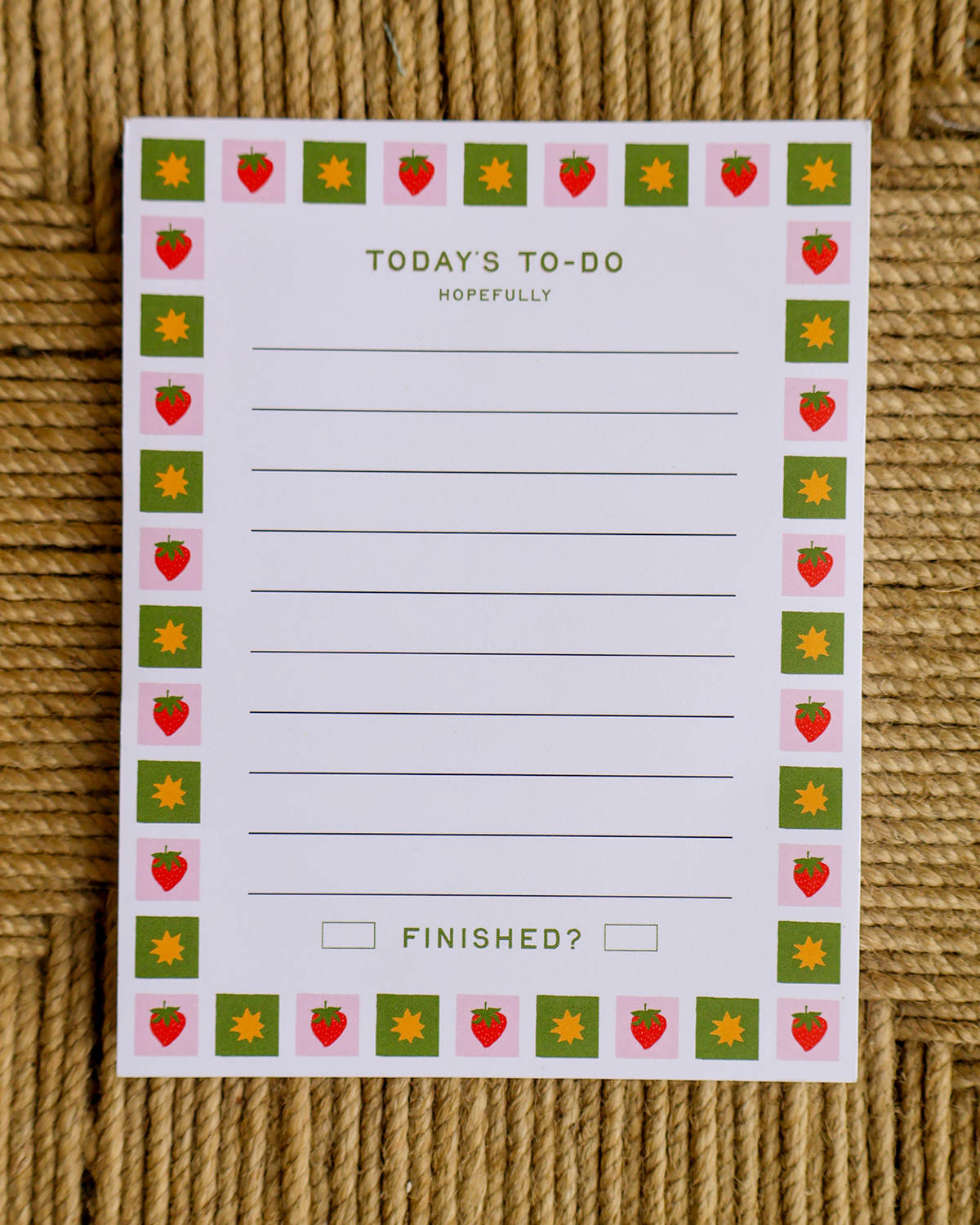 A white notepad with a decorative border of strawberries and sunflowers, titled 'TODAY'S TO-DO HOPEFULLY' and 'FINISHED?', placed on a woven tan surface.