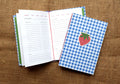 A handbound recipe log notebook with a blue and white gingham cover and a strawberry illustration on the front, shown open with recipe pages inside on a brown burlap background. 