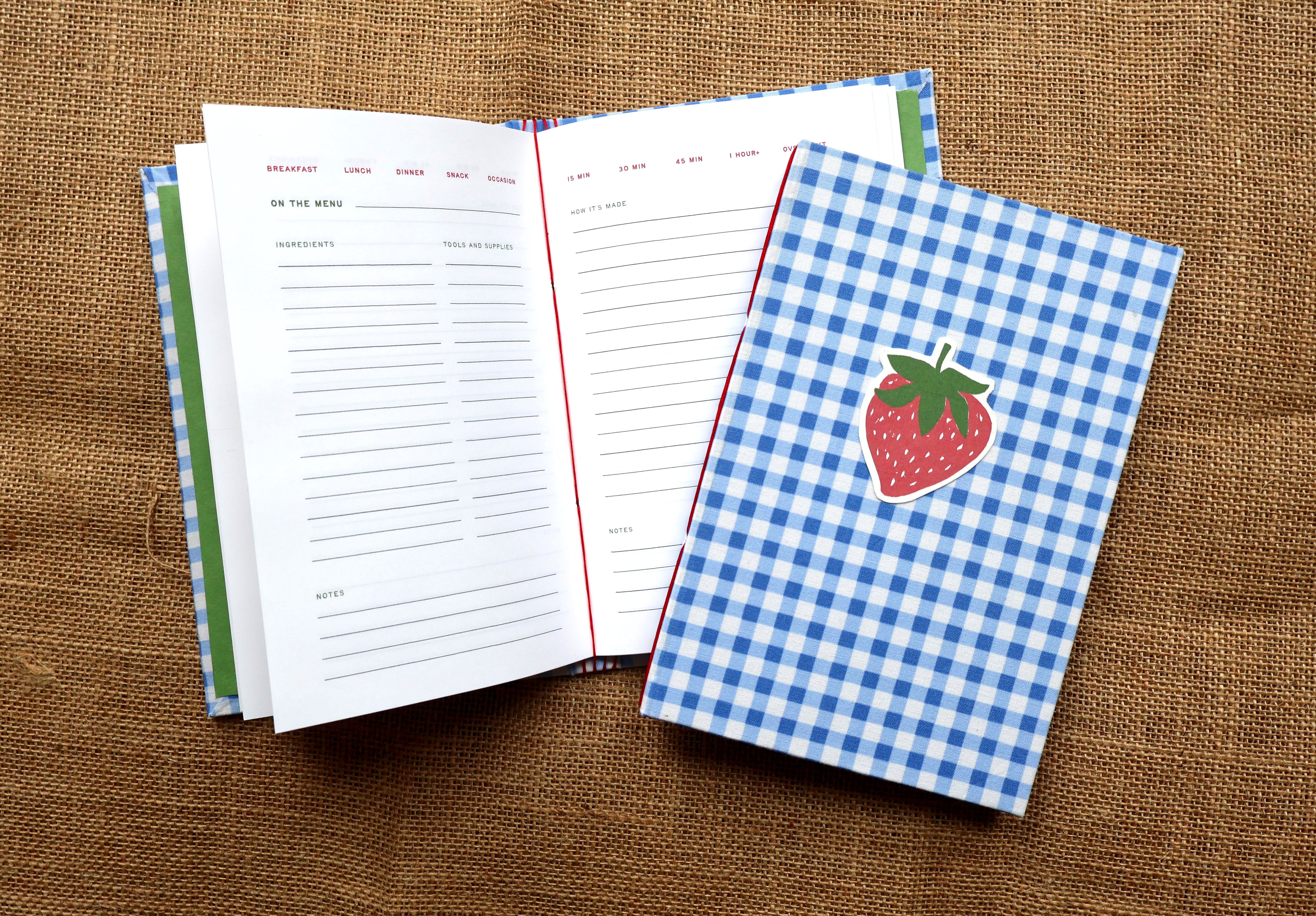 A handbound recipe log notebook with a blue and white gingham cover and a strawberry illustration on the front, shown open with recipe pages inside on a brown burlap background. 