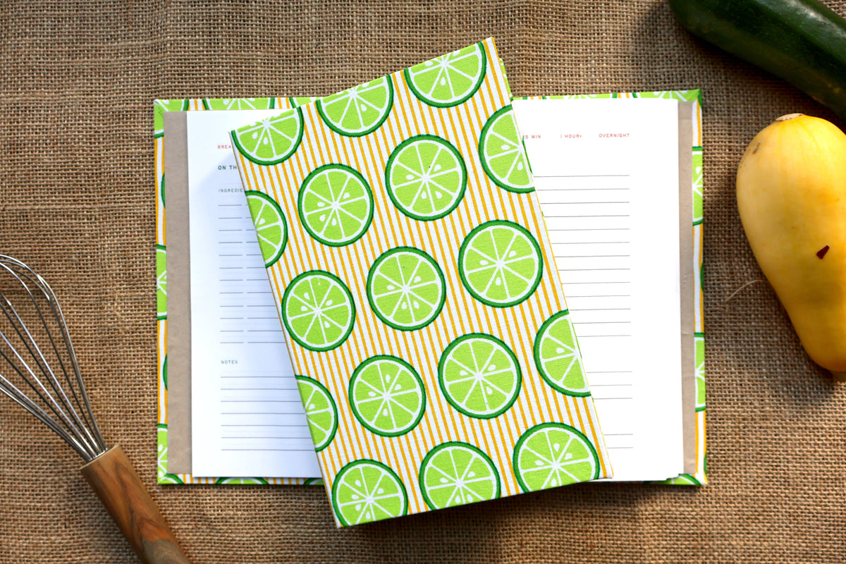 A hardcover recipe log with a green and orange lime slice patterned cover, shown open with lined pages inside, styled on a burlap surface with a whisk, cucumber, and mango nearby.