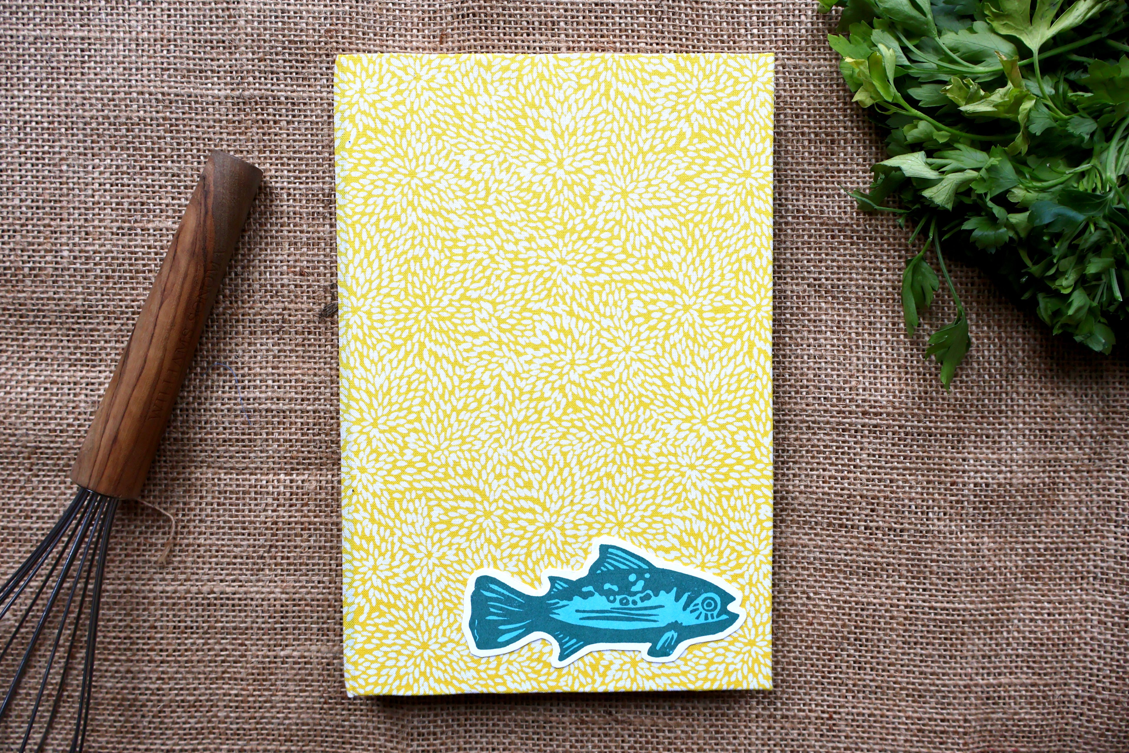 A yellow trout recipe log with a white leaf-like pattern and a blue fish linocut design at the bottom, photographed from above on a brown burlap surface with a wooden whisk on the left and green herbs on the right.