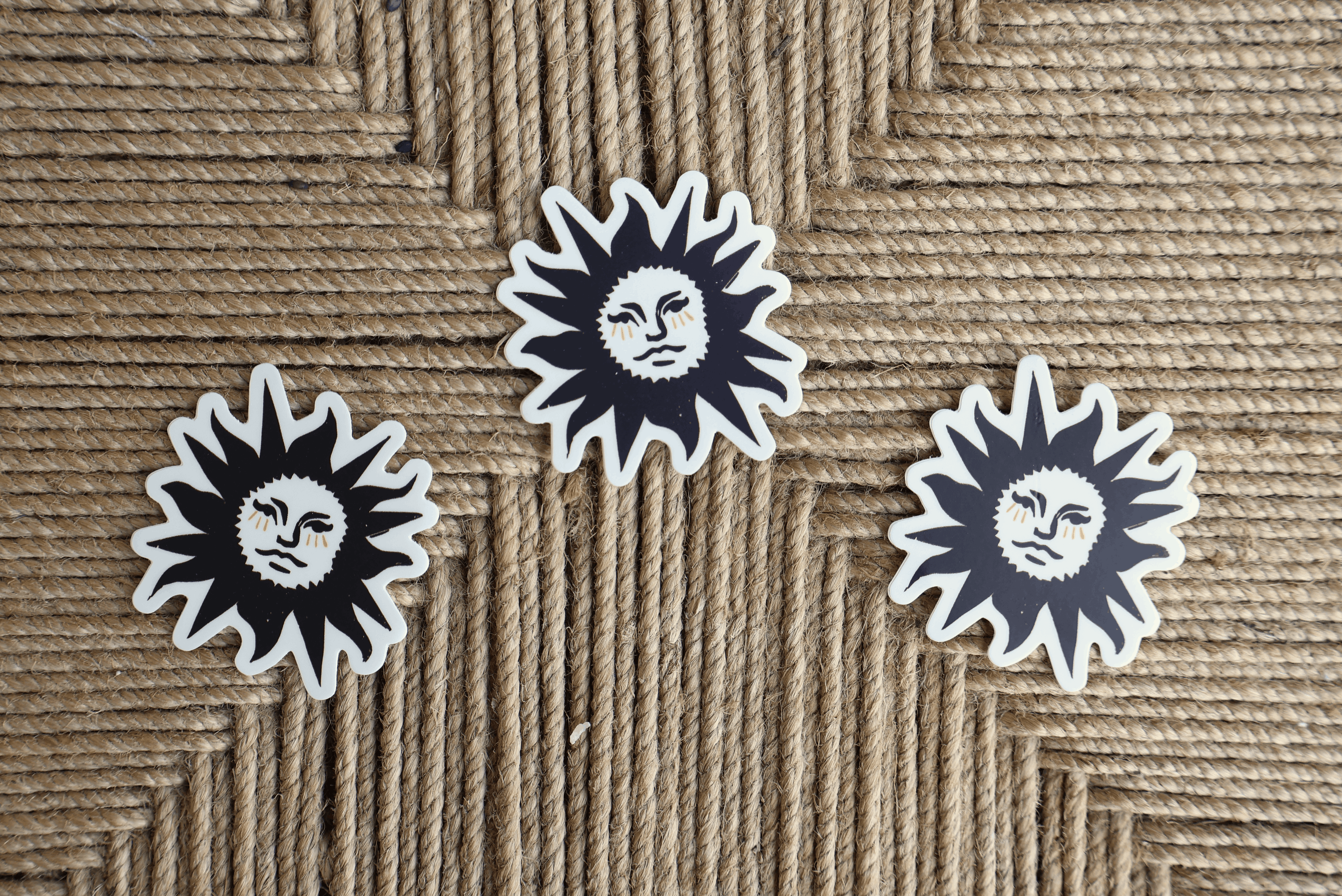 Three die-cut vinyl stickers featuring a black sun face with white rays and a white border, arranged on a woven jute rope background.