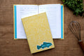 A yellow patterned hardcover recipe log with a teal fish linocut design on the cover, styled on top of an open recipe page on a burlap surface with a whisk and parsley nearby.