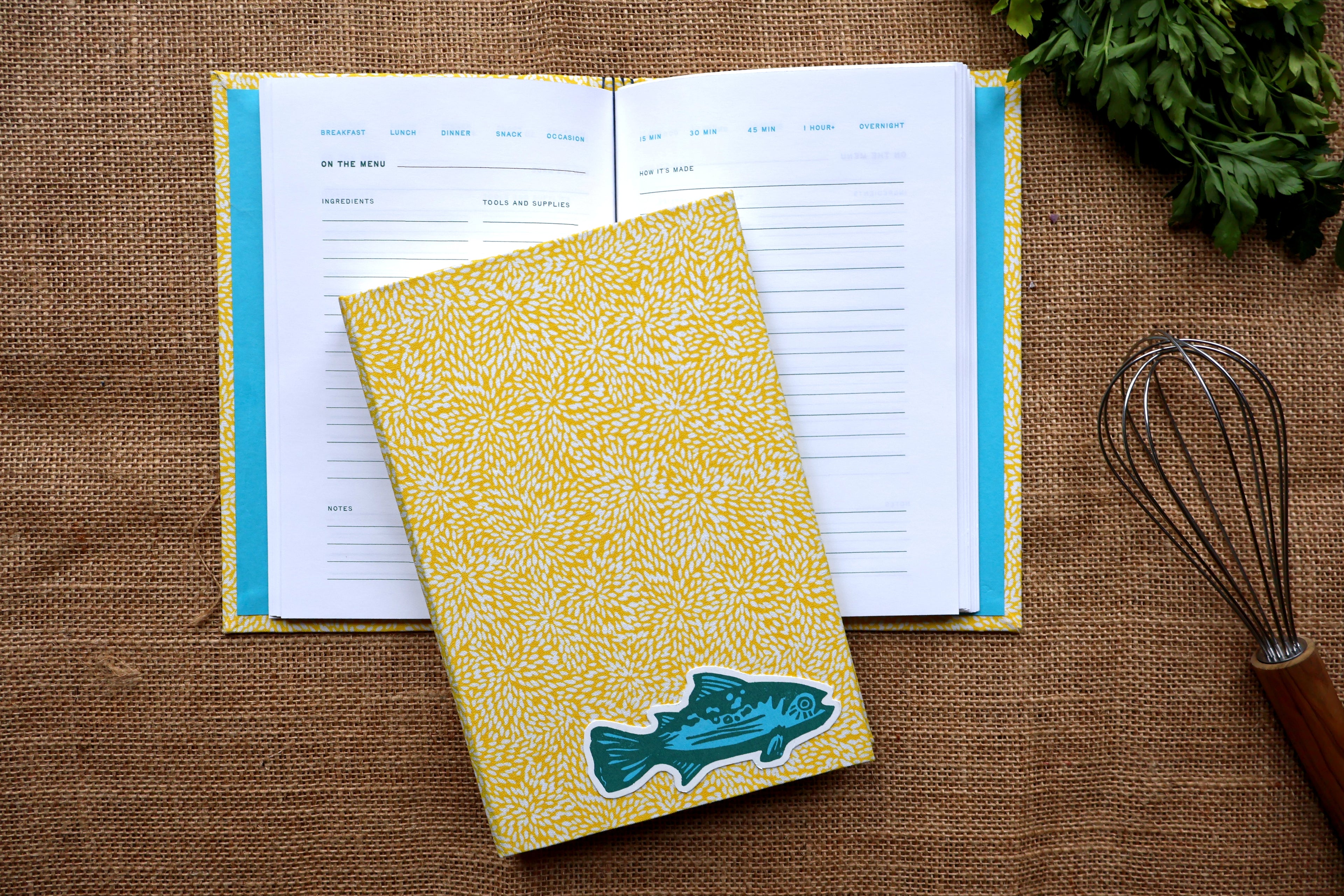 A yellow patterned hardcover recipe log with a teal fish linocut design on the cover, styled on top of an open recipe page on a burlap surface with a whisk and parsley nearby.