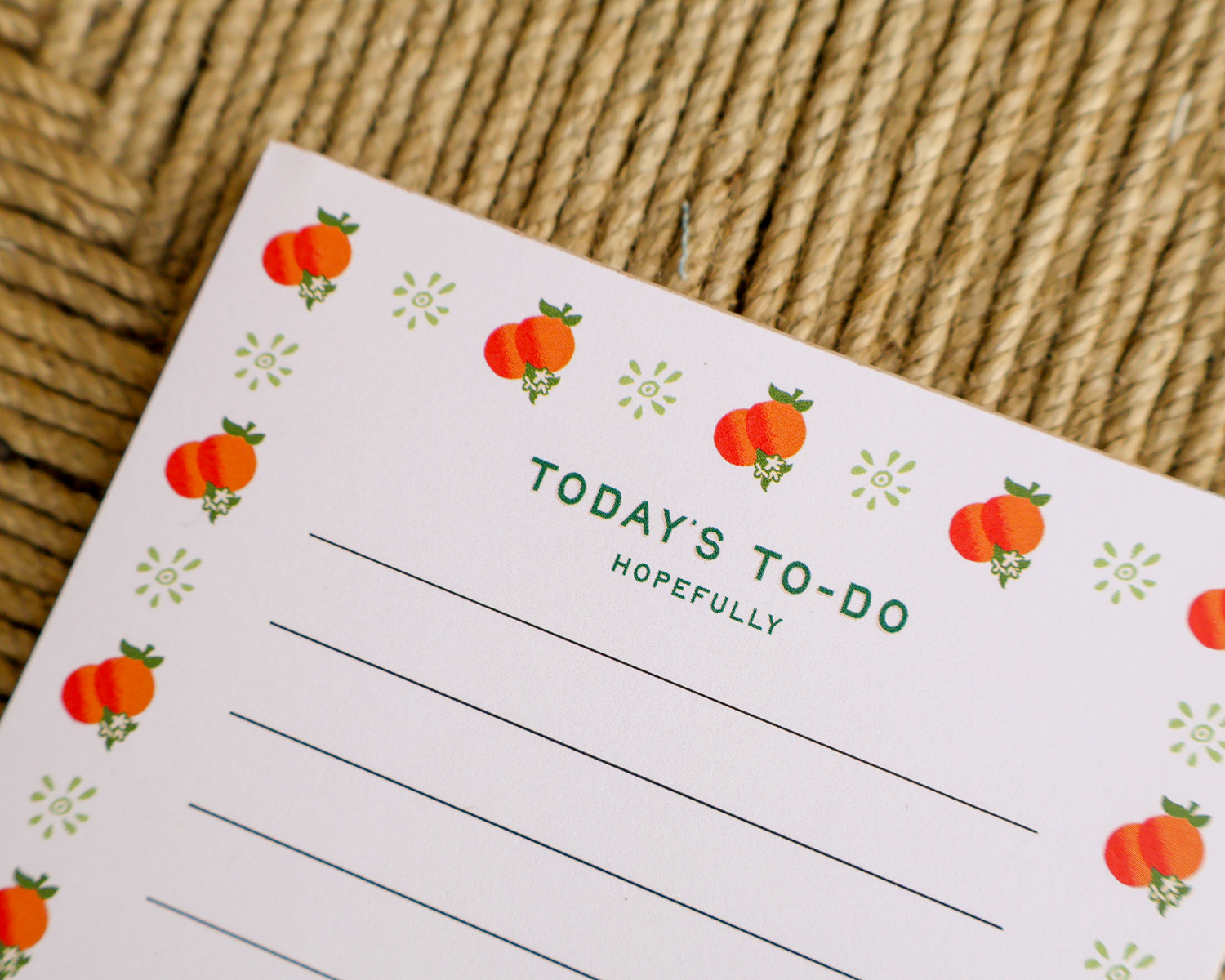 A close-up of a white to-do list notepad with green text reading 'TODAY'S TO-DO HOPEFULLY' and orange fruit illustrations along the border, placed on a woven straw surface.