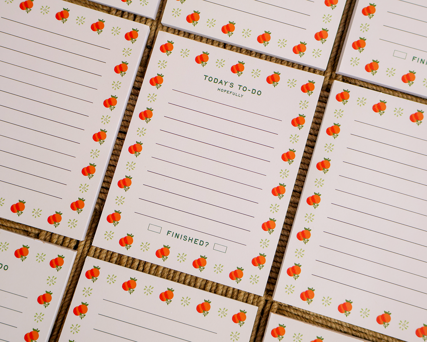 A close-up of a grid of white to-do list cards with a repeating border of oranges and small green leaves, arranged on a brown textured surface. Linocut Design