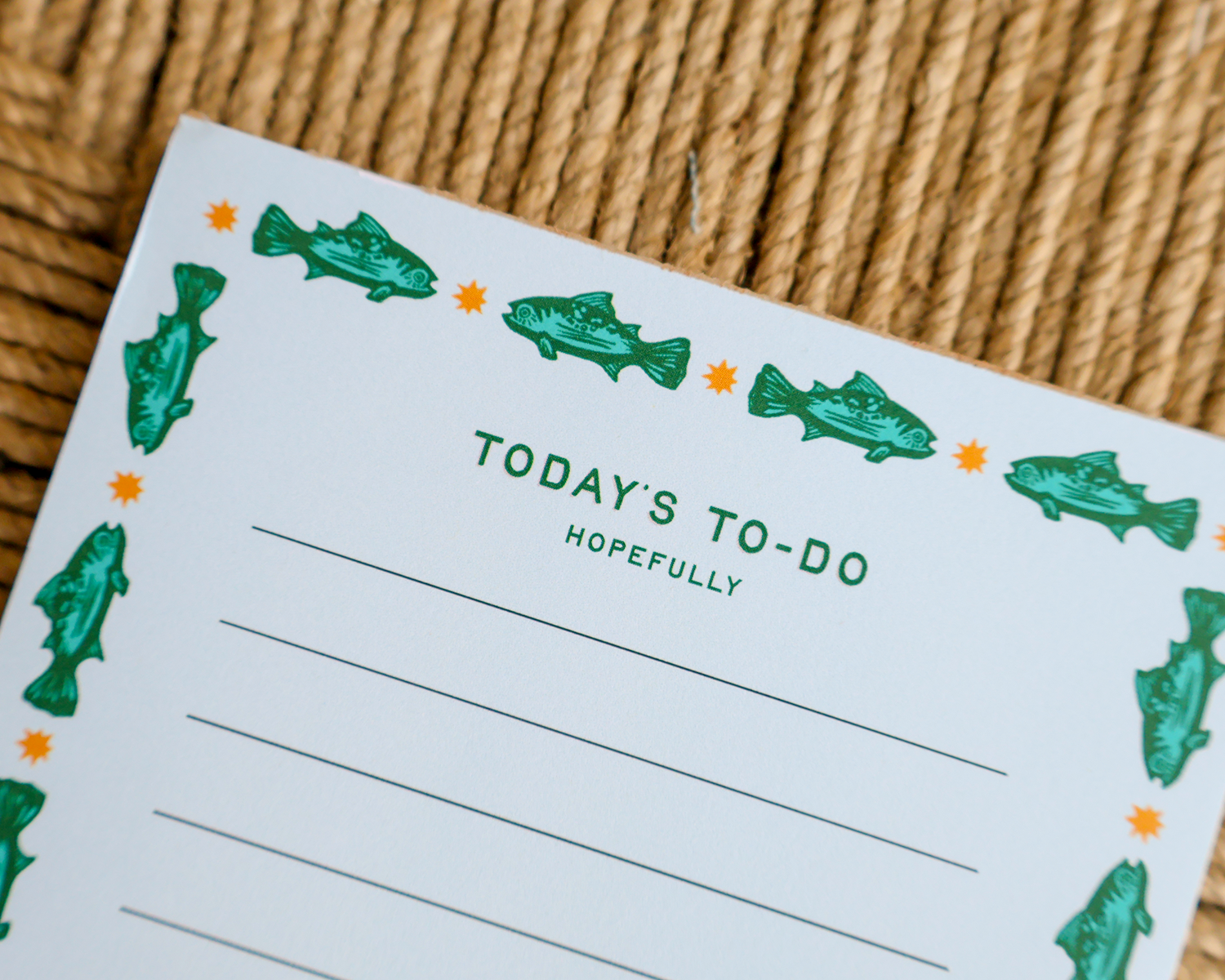 A close-up of a white notepad with green fish illustrations around the border and the text 'TODAY'S TO-DO HOPEFULLY' printed in green, placed on a woven tan rope surface.