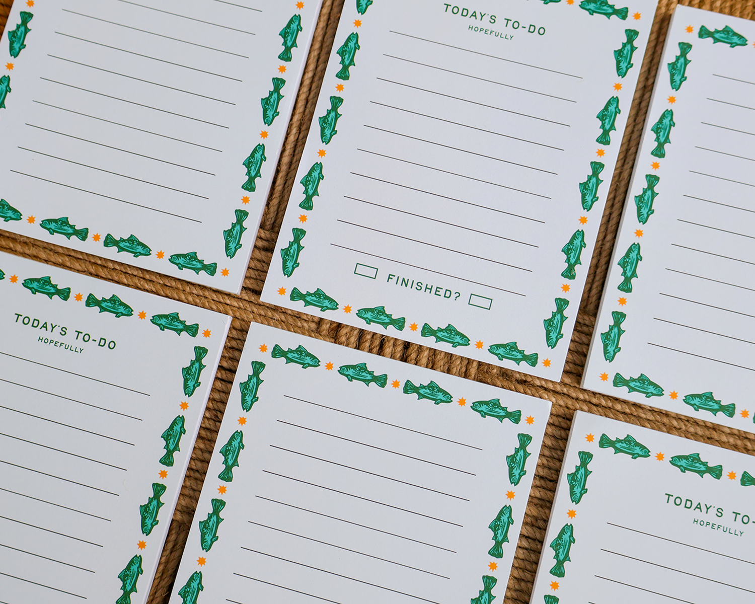 A close-up of several white to-do list cards arranged in a grid on a brown woven mat. Each card has a green fish border, orange dots, and the text 'TODAY'S TO-DO' and 'hopefully'. Linocut Design