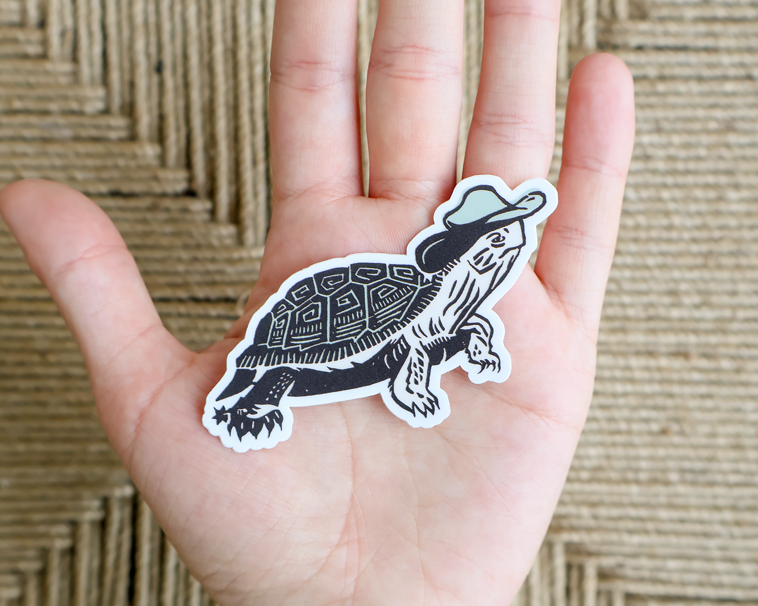 A die-cut sticker of a black and white illustrated turtle wearing a light blue hat, held on the back of a person's hand against a woven straw background. Linocut Sticker